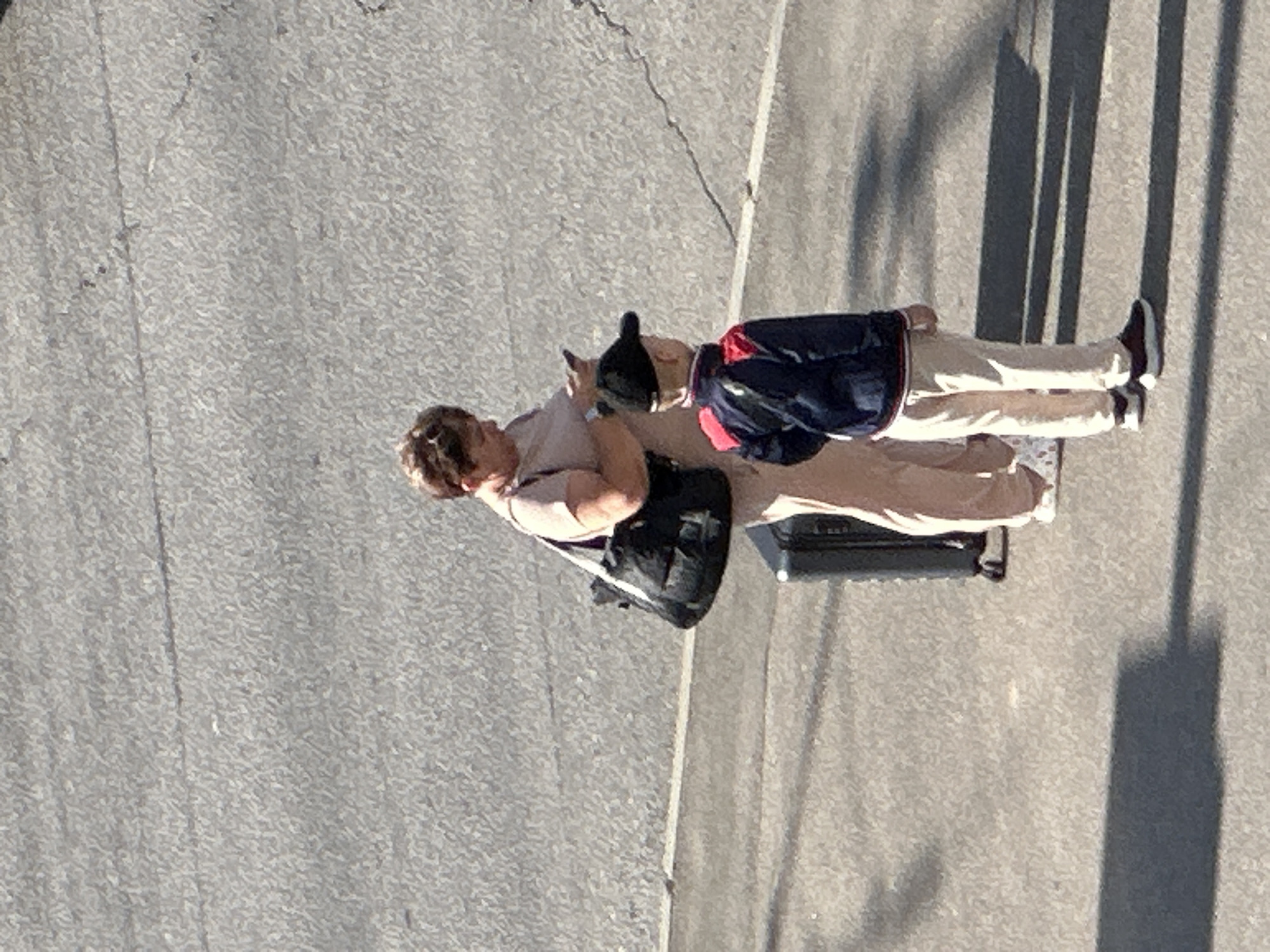 An adult woman and a young boy stand on an asphalt surface. The woman, wearing a light pink shirt and pants, is looking at her phone and holding a black handbag over her left shoulder. A wheeled suitcase is positioned behind her. The boy, wearing a navy blue jacket with red accents, tan pants, and a black baseball cap, stands facing away from the camera. The scene appears to be a street or public walkway in Podolsk, Russia, with the ground showing signs of wear and cracks. Shadows suggest a sunny day.