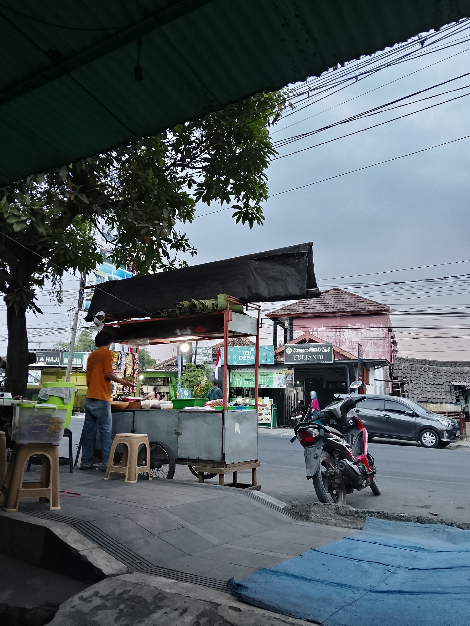 The image depicts a street-level view in Balun, Indonesia, characterized by a cloudy sky and overhead power lines. In the foreground, a food cart with a black tarp cover is set up on a sidewalk. A person wearing an orange shirt and blue jeans stands behind the cart, seemingly engaged in preparing or serving food. A red and black motorcycle is parked on the street to the right of the food cart, partially obscuring a gray sedan. The background features buildings, including one with a pink facade and a red tiled roof, bearing signage that includes "Sanggar Rina & Tari YULIANDI." Various plants and trees are also visible, contributing to the urban streetscape. The sidewalk area includes plastic chairs and containers.