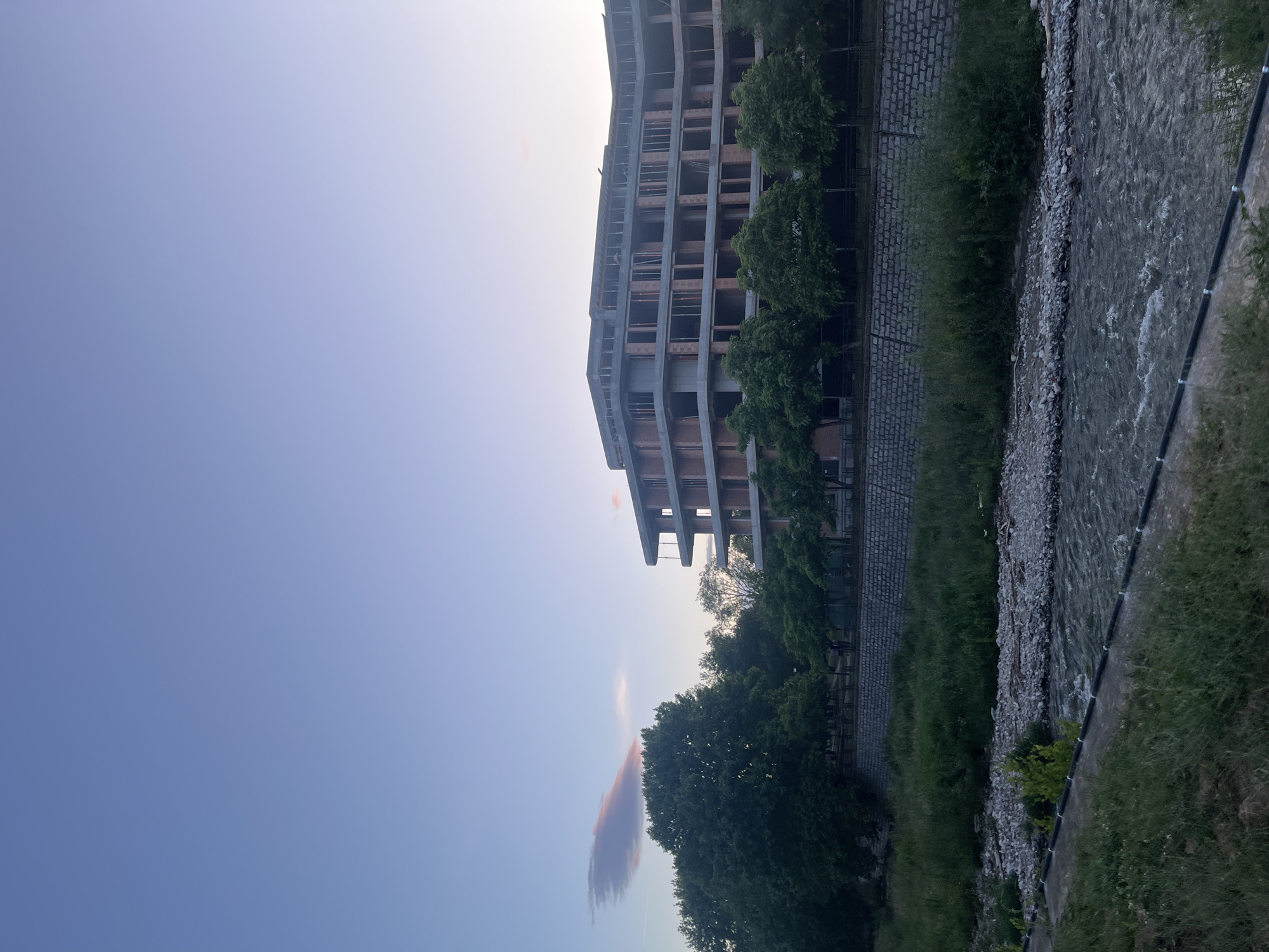 The image captures a modern, unfinished building next to a river. The building's concrete structure is visible, with repetitive window or balcony frames on each floor. Trees line the space between the building and the riverbank. The river flows from the upper right to the lower right of the frame. The sky is a pale blue, with a low-hanging, reddish-brown cloud visible below the building. This scene may be located in Blagoevgrad, Bulgaria.