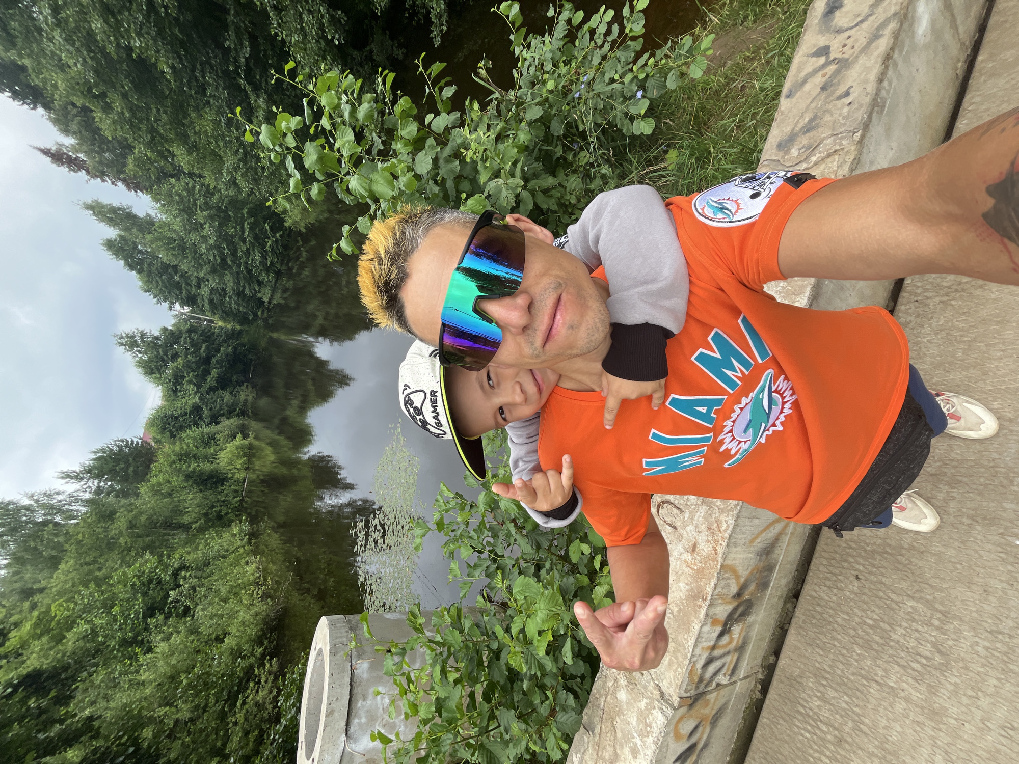 In this selfie taken outdoors, a man and a young child are posing in front of a body of water surrounded by lush green trees. The man, with dyed blonde hair and striking, iridescent sunglasses, has his arm around the child who is perched on his shoulders. Both are wearing orange t-shirts with a dolphin logo and the word "MIAMI" printed on them. The man is making a "rock on" hand gesture, while the child points towards the camera. The background shows a reflection of the trees in the calm water, suggesting it is a clear day. A concrete structure, possibly a bridge or embankment, runs along the foreground, with graffiti visible on its surface. The overall mood appears cheerful and playful, capturing a moment of bonding between the two subjects. The text "GAMER" is visible on the child's cap.