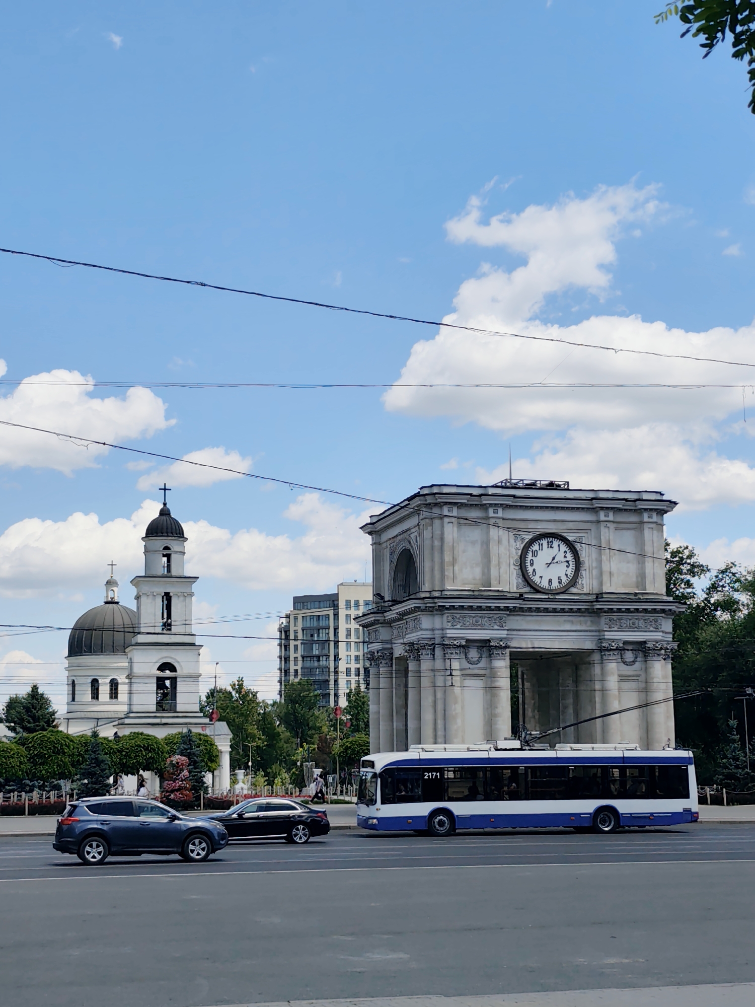 This is a daytime, eye-level, full shot of a city street scene in Chișinău, Moldova. The foreground features a grey asphalt road with two lanes of traffic. On the left lane, a dark grey SUV is driving towards the viewer, and in the right lane, a black sedan is also driving towards the viewer. In the background, a blue and white trolleybus is traveling away from the viewer on the right side of the road. To the left of the trolleybus is the Triumphal Arch, a large stone monument with a prominent clock face. Behind the arch, a white cathedral with a dark dome and a bell tower topped with a cross is visible. Further back, a modern high-rise building stands tall against the sky. The sky is bright blue with scattered white clouds. Overhead, thick black wires crisscross the frame, likely power lines for the trolleybus. Green trees and decorative bushes line the street.
