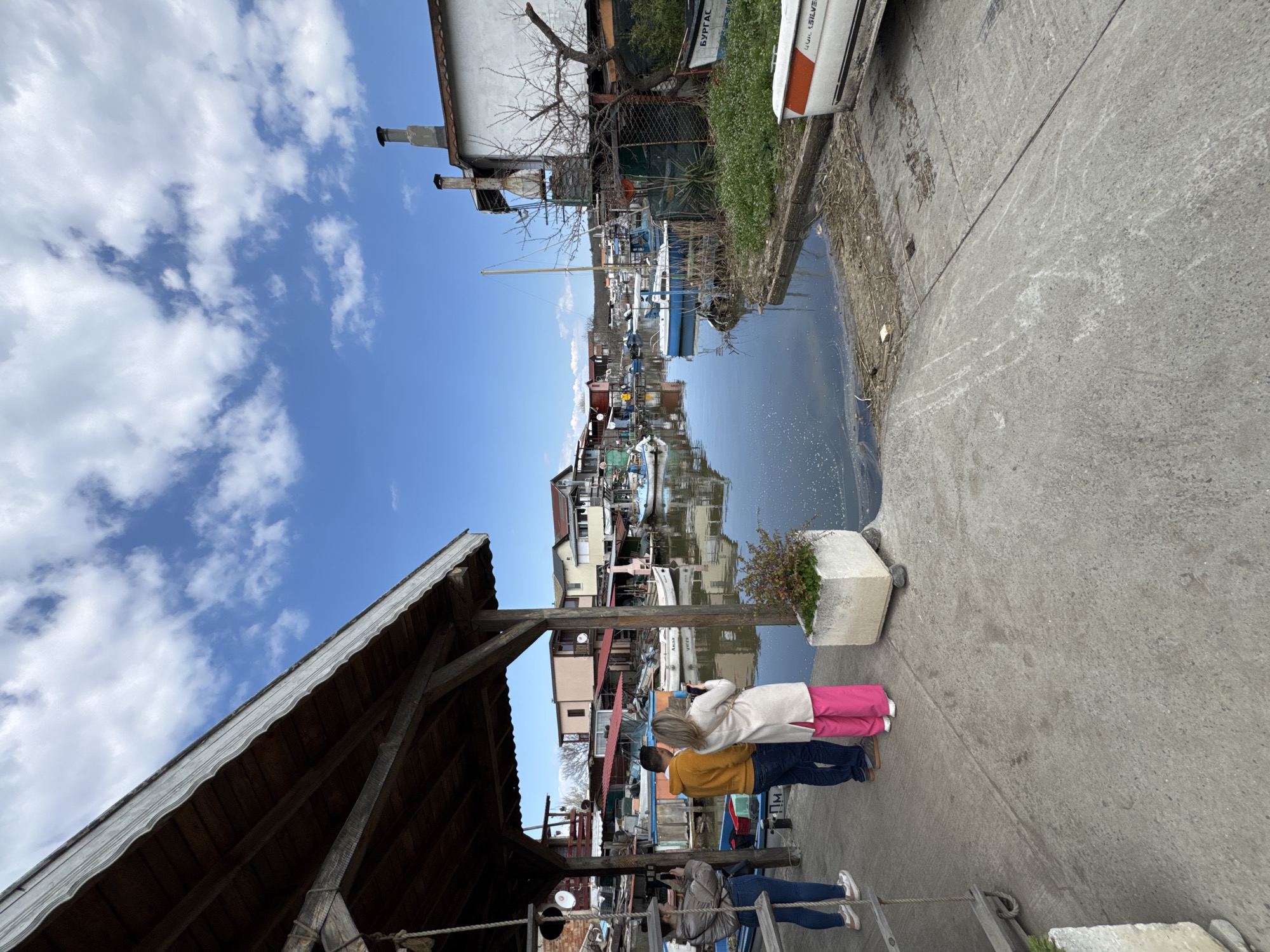 The image depicts a canal scene in Burgas, Bulgaria. Two people, one in a yellow jacket and the other in a white coat and pink pants, stand on a concrete quay, observing the canal. Reflections of buildings and boats are visible in the water. A woman in a grey jacket is taking a photo. A wooden structure with a sloped roof is on the left. Vegetation and a partially visible white car are to the right, beyond the canal. The sky is blue with clouds.