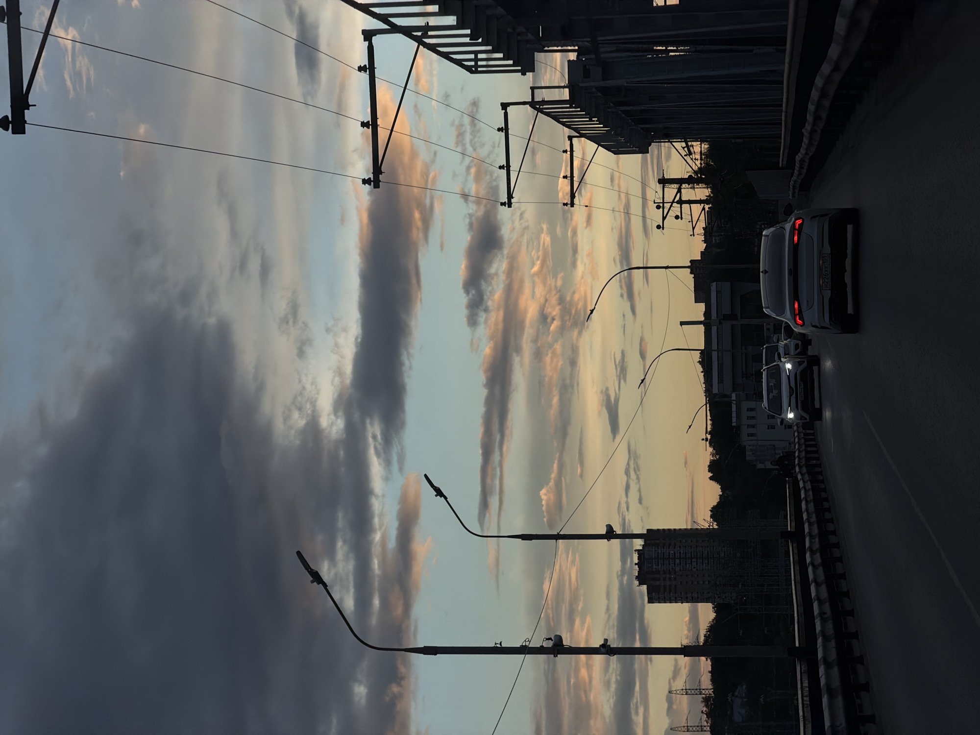 The image captures a view from a bridge in Perm, Russia. The sky is filled with dramatic clouds, reflecting the colors of a setting sun. Below the bridge, city buildings are partially visible. Two cars are on the bridge, heading away from the viewer. Streetlights line the sides of the road. Infrastructure elements like support structures and power lines are also visible, cutting across the sky.