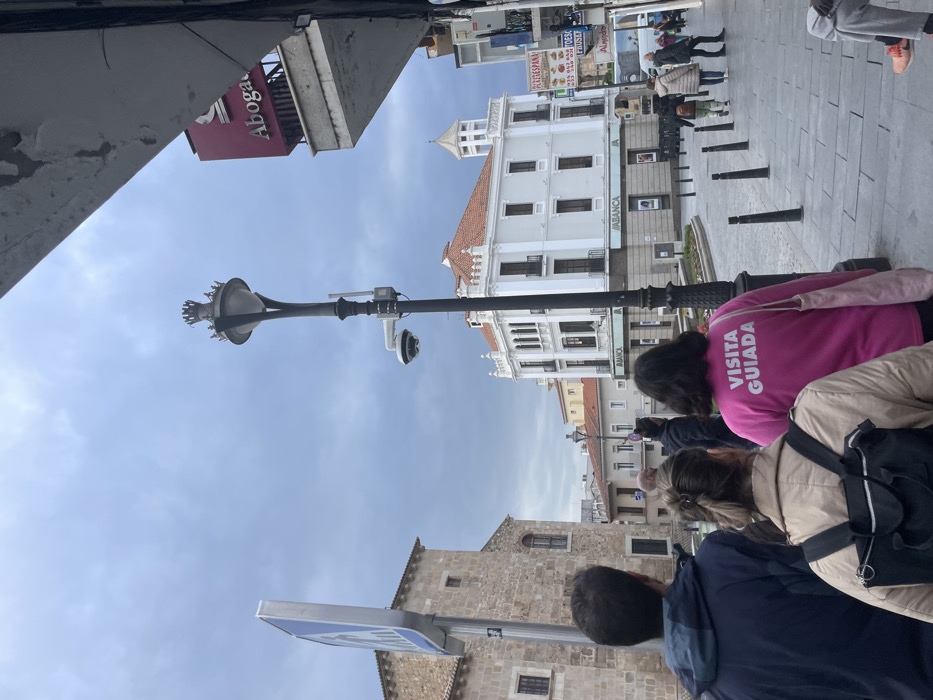This media file captures an outdoor urban scene in Mérida, Spain, likely a street or plaza, under an overcast sky during daytime. The perspective is slightly angled upwards, looking past foreground elements towards buildings in the mid-ground and background.
In the immediate foreground, facing away from the viewer, are several individuals. One prominent person wears a bright pink long-sleeved top with the white text "VISITA GUIADA" (Guided Tour) printed across the back, strongly suggesting they are a guide or part of a tour group. Another person wears a light beige jacket and a black backpack.
To the left, a tall, ornate black street lamp with a distinctive, flared top stands vertically. Attached to this lamppost, below the main light fixture, is a white, dome-shaped surveillance camera. Partial views of architectural structures, including a concrete overhang, frame the upper left and lower left of the image. A red sign with "ABOGA" (likely part of "Abogacía" or "Abogado" - Law/Lawyer) is visible on a building on the upper left.
The mid-ground features a large, ornate white building with multiple windows and a tiled, reddish-brown roof, possibly a historic structure. The name "ABANCA," a Spanish bank, is visible on its facade. The ground is paved with light-colored stones, and several black bollards line the edge of the street or pedestrian area. In the background, more people are visible, some walking, and additional buildings of varying architectural styles can be seen under the expansive, light blue-grey sky. The overall atmosphere appears calm, indicative of a typical day in a city center.