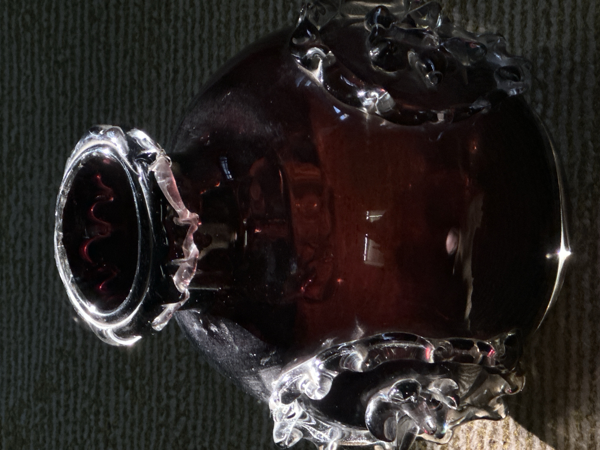 The image shows a close-up, vertical, full-body shot of a dark red glass bottle with clear glass decorative elements on its sides. The bottle has a ruffled rim and the glass is highly reflective, with highlights and shadows emphasizing its curves and texture. The background is a textured, dark surface, possibly a rug or fabric, with diagonal lines of light and shadow suggesting a light source from the left. There are no people or actions depicted. The image does not provide any information about its location in the city of Luhansk, Ukraine.