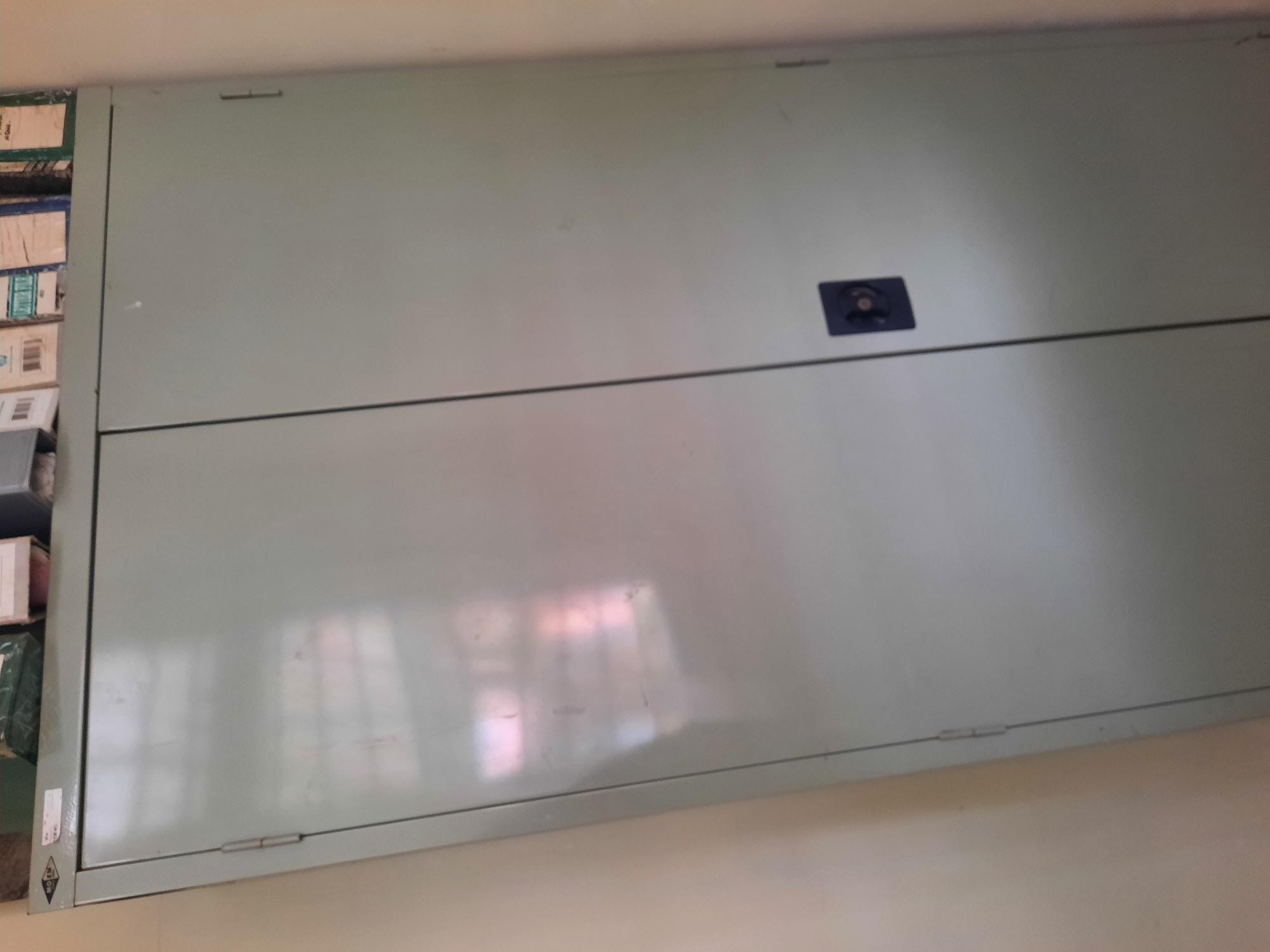 The image shows a closed, light gray metal cabinet with two doors, set against a light beige wall. The cabinet has a dark gray handle on the right door. A reflection of what appears to be a window with multiple panes is visible on the left door of the cabinet. The top of the cabinet is partially obscured by stacked items, including cardboard boxes and what looks like a green item. A label with text is visible on the top left edge of the cabinet. The scene appears to be indoors, possibly in an office or storage area. The specific location in Lhokseumawe, Indonesia cannot be determined from the visual information provided.