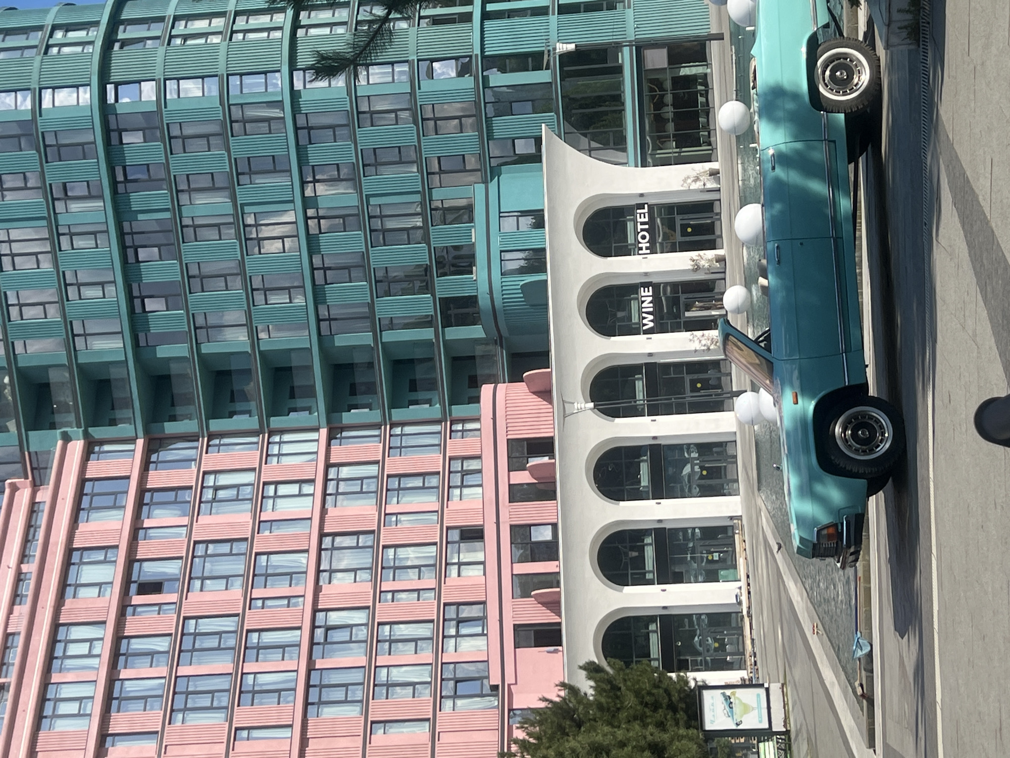 The image shows a modern hotel complex in Odesa, Ukraine, featuring two brightly colored buildings: one pink and one teal. The teal building is curved and has a pattern of vertical lines. The pink building is rectangular and also has vertical detailing. Both buildings have numerous windows with sheer white curtains visible.
In front of the buildings, there is a white structure with a series of arched entrances, one of which displays the text "WINE HOTEL". The area in front of the hotel appears to be a public space with a water feature or shallow pool.
A vintage teal convertible car is parked in the foreground, positioned to the left of the water feature. The car has a chrome trim and white-walled tires. Several large white spheres are visible in and around the water feature, and one is behind the car.
To the left of the scene, there is a sidewalk with a bench and a signpost displaying an image of a cocktail and a vehicle. A tree is visible on the far left. The ground is paved with grey and white patterned tiles. The sky is visible at the top of the image, appearing bright and sunny. The overall impression is one of a contemporary urban environment with a retro touch from the car.