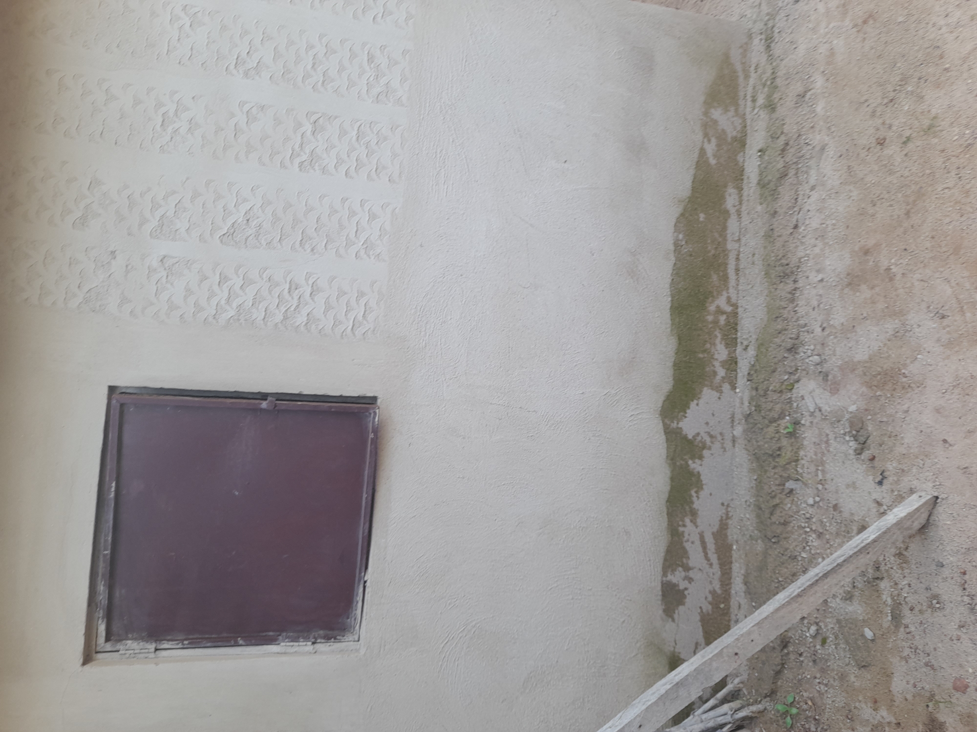 An exterior shot of a light-colored building wall in Ibi, Nigeria, shows a dark-colored, rectangular metal window with a visible hinge on the left side. The wall above the window features a textured pattern of raised, repeating wave-like shapes arranged in vertical columns. Below the window, the wall transitions to a rougher texture. At the base of the wall, a band of dampness with green algae growth is visible against the sandy ground, which is littered with small stones and a few blades of grass. A portion of a wooden plank, leaning diagonally from the lower left corner into the frame, is also present on the ground. The overall impression is of an unfinished or weathered exterior wall.