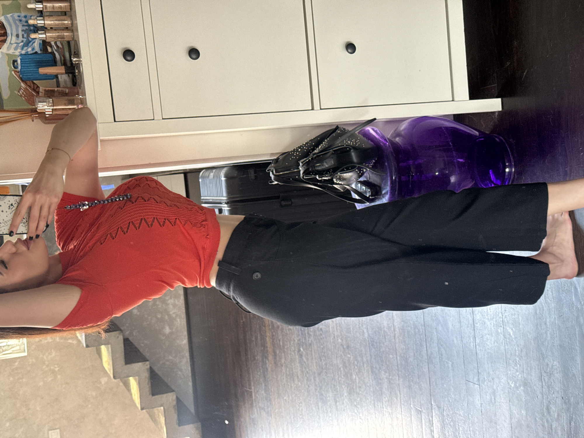 A young woman is laying down on a wooden floor indoors. She is wearing a red cropped top and black pants. Next to her are a purple glass vase, a black studded purse, and a black suitcase. Behind the woman is a white cabinet with dark knobs. There are stairs and a wall decorated with drawings in the background. The location cannot be determined to be in Sofia, Bulgaria from the image alone.