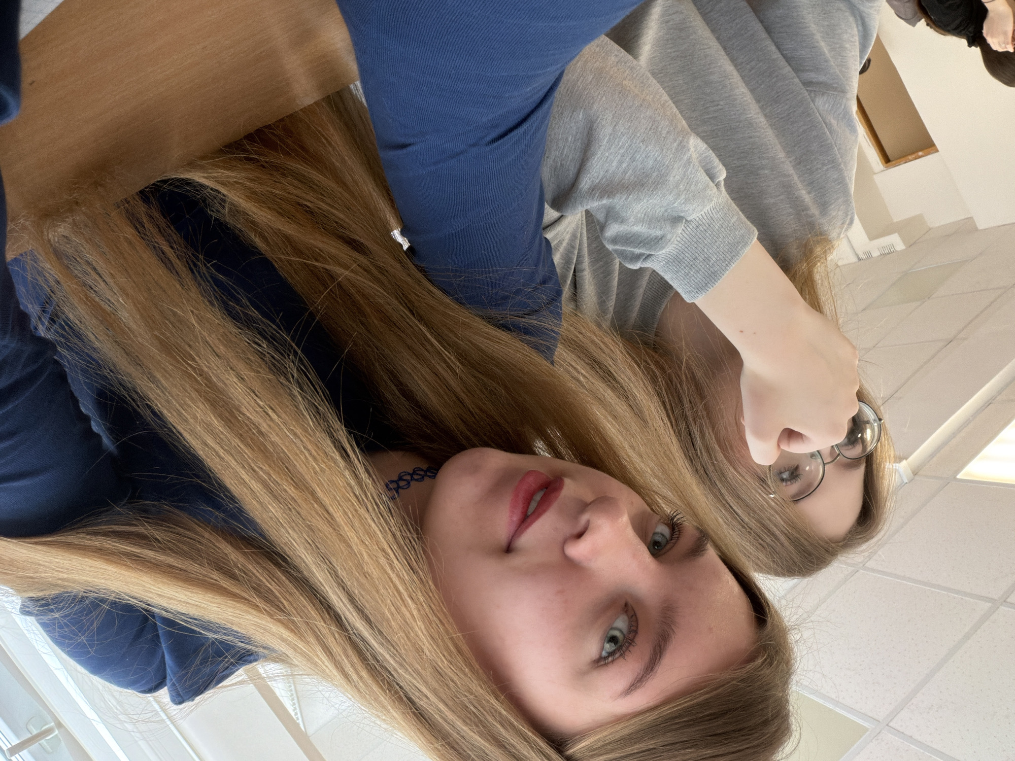 The image depicts the interior of an institutional or educational building in Chaikovsky, Russia. Two young adult females are shown from a low-angle, selfie-style perspective.
In the foreground and to the right, a female with long, straight, light brown hair, light-colored eyes, and red lipstick is visible. She wears a dark blue long-sleeved top and a dark blue choker necklace. Her head is tilted slightly, and she looks directly at the camera with a subtle smile. Her right arm extends towards the bottom center of the frame, likely holding the capture device.
To the left and slightly behind the first individual, a second female is partially visible. She has long, straight, light brown hair and wears round-framed glasses. She is dressed in a light grey long-sleeved sweatshirt. Her right hand is raised to her face, with fingers positioned near her glasses. Her gaze is directed away from the camera, towards her left.
The environment includes a white false ceiling composed of square acoustic panels and visible rectangular fluorescent light fixtures. A light-colored wall is present in the background on the left, with a partial view of a wooden door frame or opening. A light brown wooden surface, consistent with a desk, is visible in the lower right corner. The scene is illuminated by overhead artificial lighting.