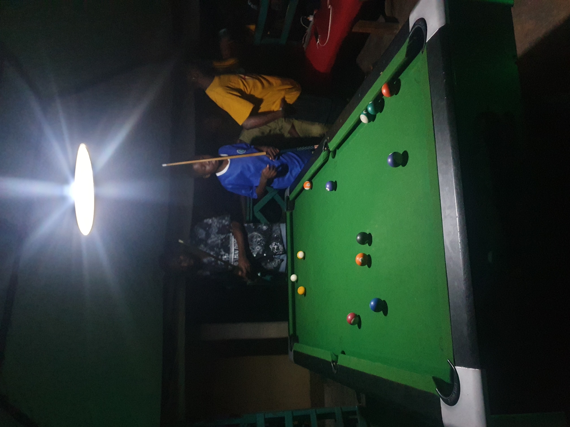 In Lokoja, Nigeria, at night, three individuals are gathered around a green pool table. The room is dimly lit by a bright overhead lamp that casts lens flare. The pool table is set up with several balls, including a white cue ball, red, blue, orange, and black balls, and some striped balls. One person, wearing a blue shirt with white trim, stands on the left side of the table, holding a pool cue. Another person, in a dark patterned shirt, stands behind them, also appearing to be playing. A third person, in a yellow t-shirt, is visible on the right side of the table, leaning over. The environment appears to be an indoor recreational space, possibly a bar or game room, with dark walls and some green metal structures visible in the background.