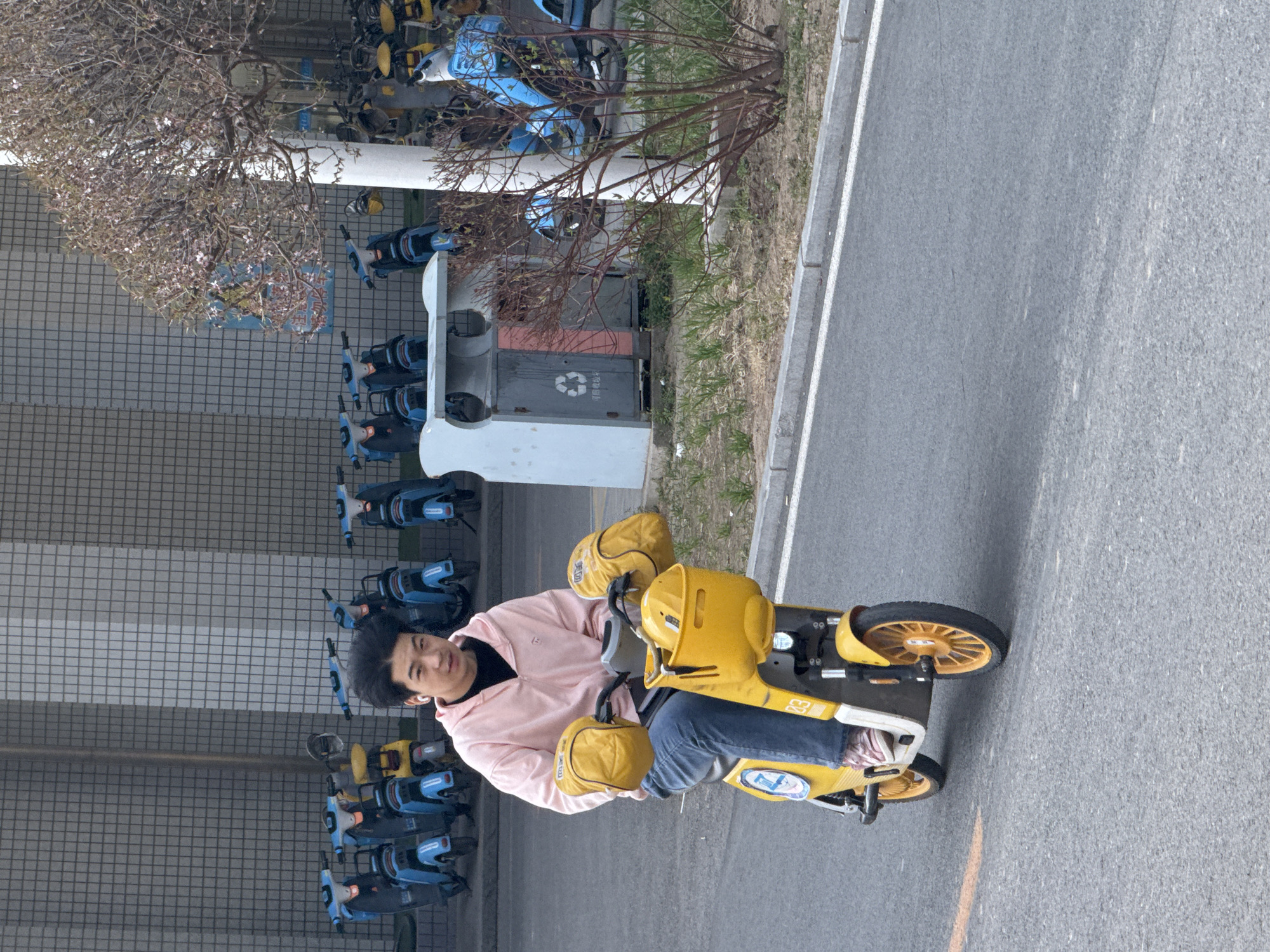 A person is seated on a yellow electric tricycle on a gray asphalt surface. The individual is wearing a light pink hooded sweatshirt, jeans, and appears to be looking towards the camera. The tricycle has yellow storage compartments on the front and handlebars. Rows of parked blue and black scooters can be seen on the left side, suggesting a bike-sharing station. There is a white and gray structure with a recycling symbol behind the scooters. A tree with bare branches is visible above the structure and the scooters, indicating it is likely springtime. The setting appears to be a street or pathway within the city of Zhongtun, China.