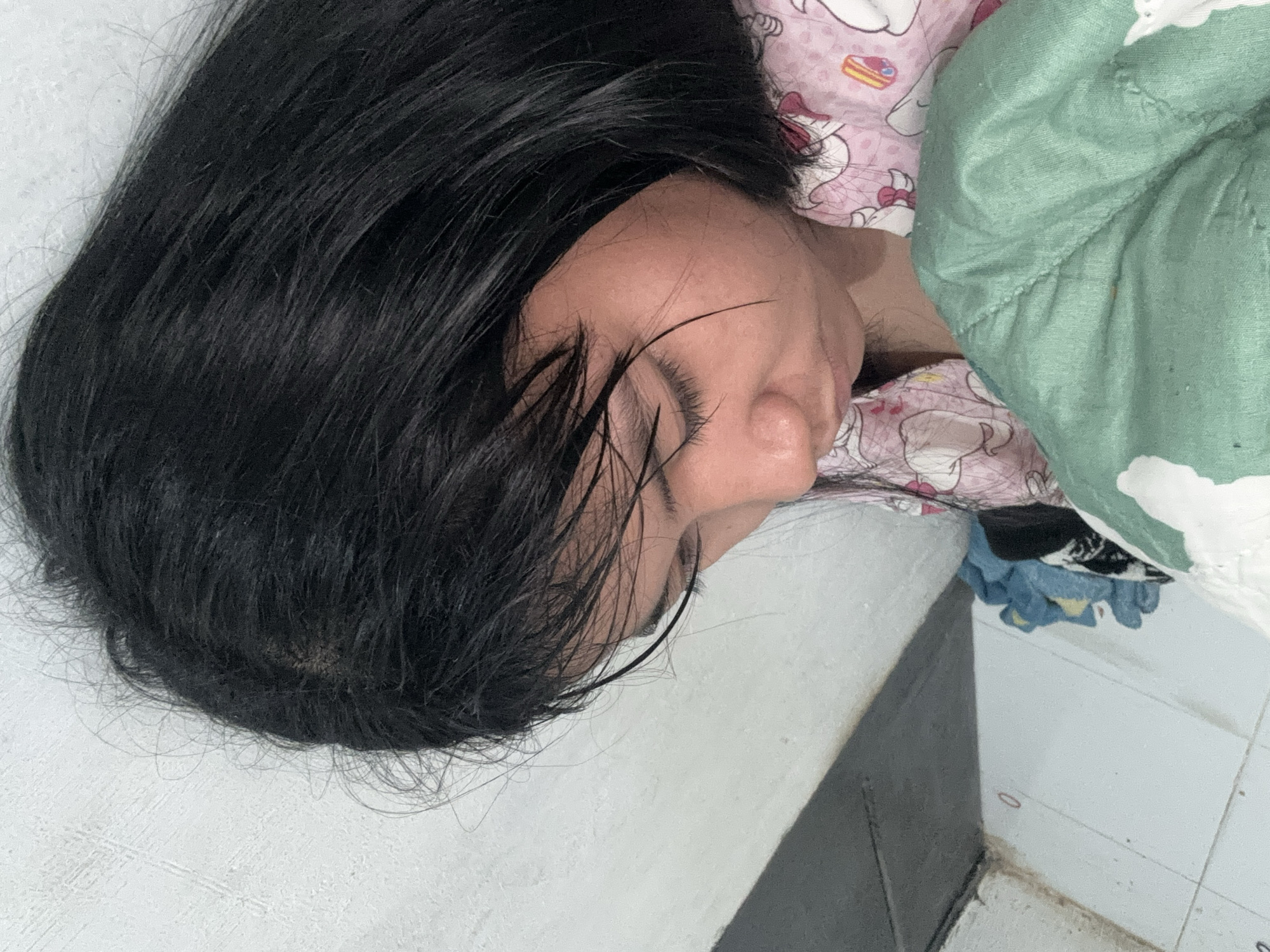 The image is a close-up shot of a young woman with black hair, looking down. She is wearing a pink pajama top with cartoon characters on it. A green blanket is draped over her chest. The background appears to be a wall with a tiled floor and some folded clothes. The scene is likely indoors, possibly in a bedroom, and the lighting suggests it could be daytime. No specific landmarks or indicators place this image in North Jakarta, Indonesia, although the style of the pajamas and the overall setting are consistent with residential areas in the region.