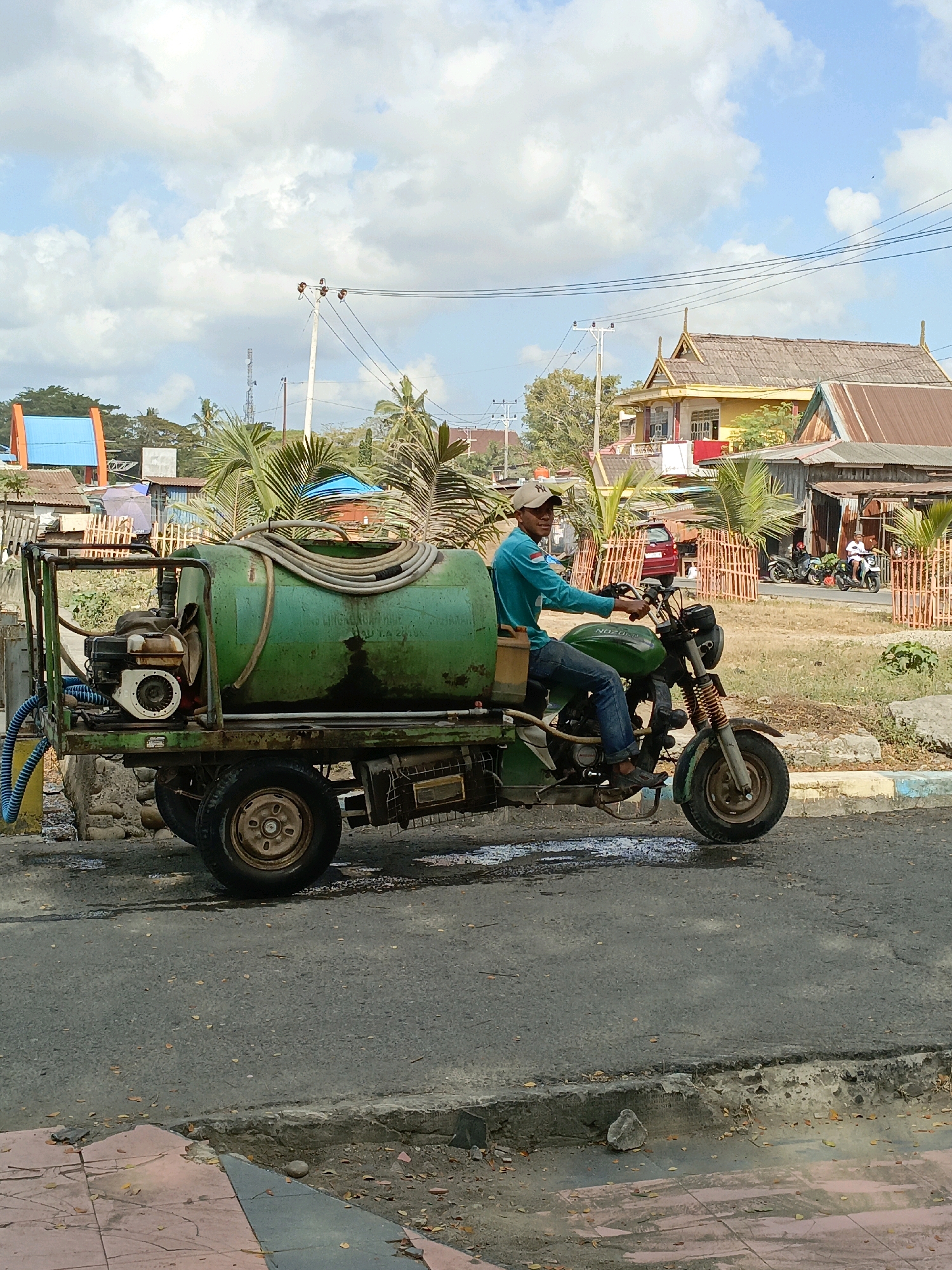 An outdoor, eye-level, medium shot captures a man on a motorized tricycle in Bulukumba, Indonesia. The man, wearing a blue long-sleeved shirt, blue jeans, and a light-colored baseball cap, is operating the vehicle which is equipped with a large green water tank, a smaller engine, and a hose. The tricycle has two wheels in the back and one in the front, and it is positioned on an asphalt road with a visible curb and sidewalk. In the background, there are several buildings with rustic, weathered roofs, palm trees, and a bright blue sky with scattered white clouds. A red car is parked behind an orange fence, and two other motorcycles with riders are also visible on the road further in the background. The overall scene depicts a typical day in a tropical urban environment.