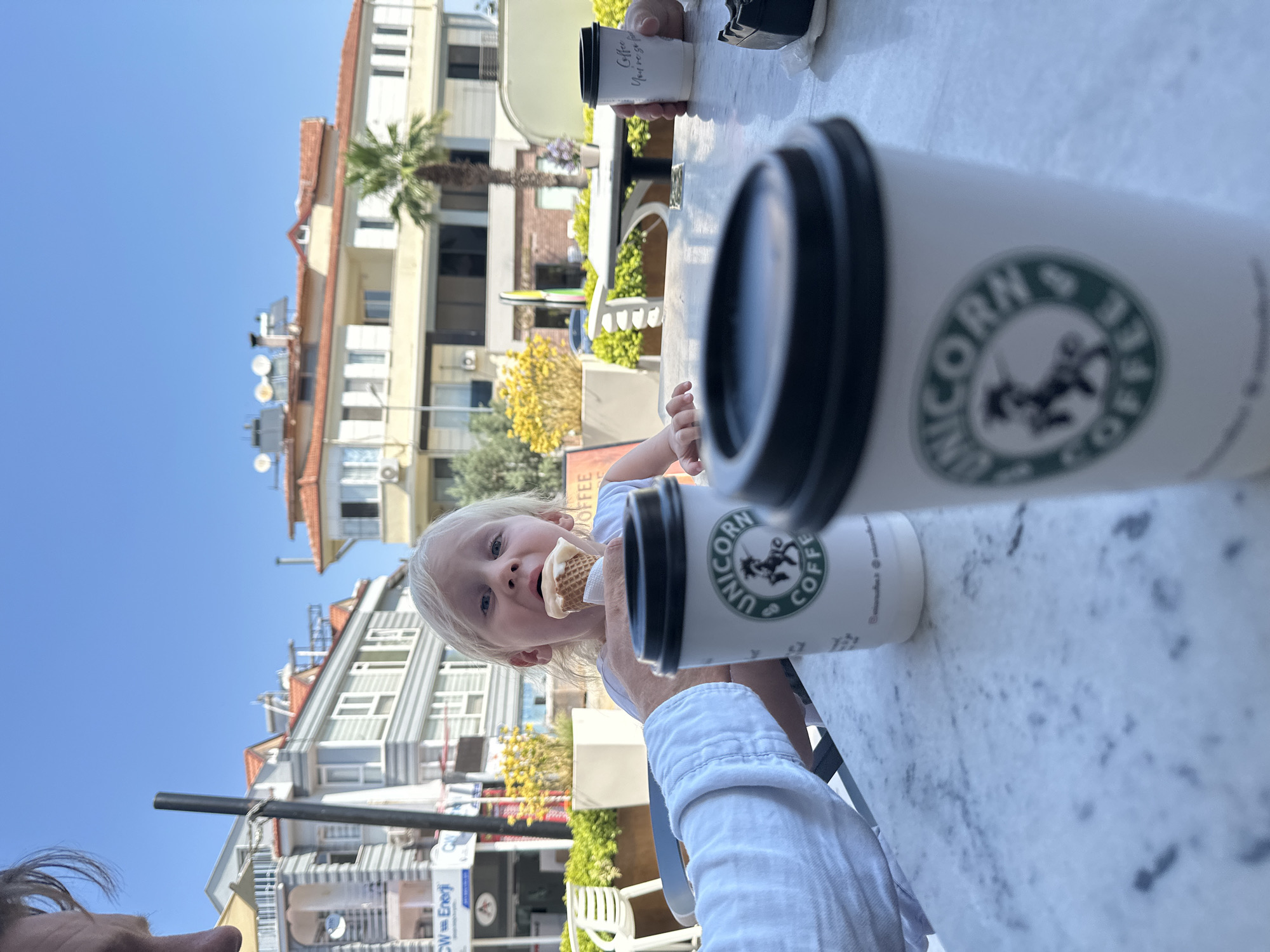 A blonde-haired toddler is seated at a marble-topped table outdoors in Fethiye, Turkey, eating an ice cream cone. The toddler is looking at the camera with a wide-eyed expression. In the foreground, there are two white paper coffee cups with black lids and a green logo that reads "UNICORN COFFEE." One cup is closer to the camera and slightly out of focus, while the other is in focus and to the left. A person's arm, wearing a white shirt, is visible on the left side of the frame, reaching towards the table. In the background, there are buildings with red-tiled roofs and white facades, typical of Fethiye's architecture. Yellow flowering bushes and a palm tree are also visible, suggesting a warm climate. The sky is clear and blue. The overall scene depicts a casual outdoor cafe setting.