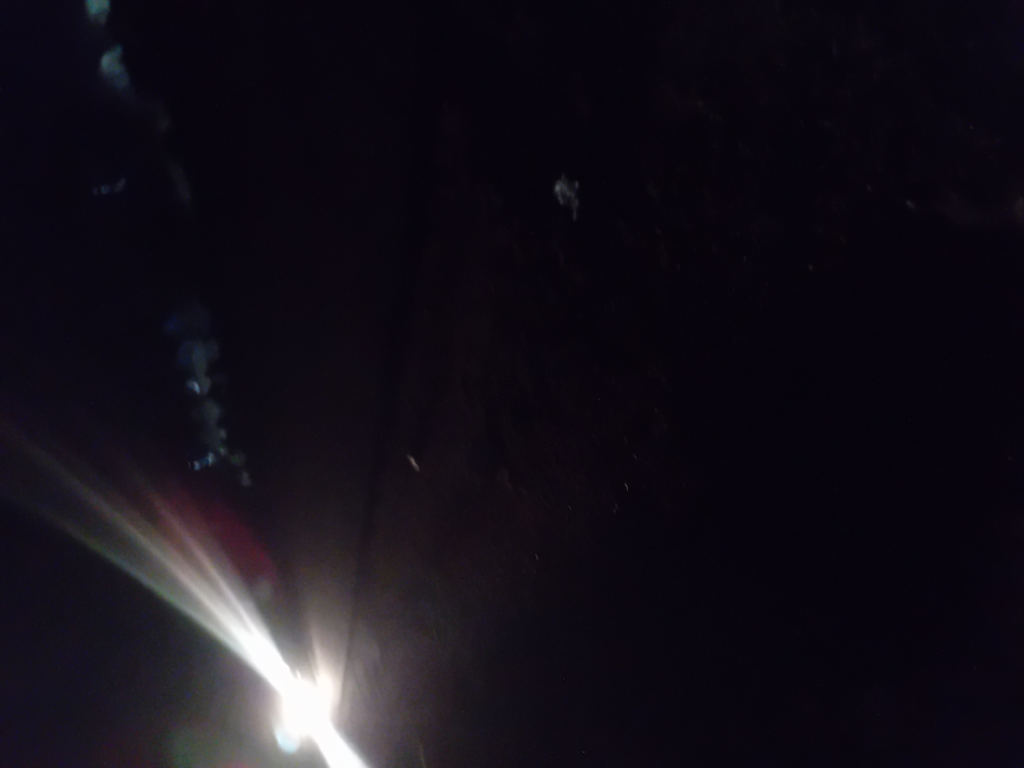 The image depicts a dark outdoor scene, likely at night, in Jauro Yinu, Nigeria. A bright light source, possibly headlights from a vehicle, illuminates the left side of the frame with a strong beam extending upwards and to the right. The light creates lens flares and streaks of white and pink. The ground appears to be a dark, possibly asphalt or dirt surface. In the background, a faint line of small, blurry lights suggest the presence of distant structures or other vehicles. Small, indistinct specks are visible throughout the dark areas of the image, which could be dust, insects, or other airborne particles. There are no clearly identifiable people or specific objects in the foreground or midground.