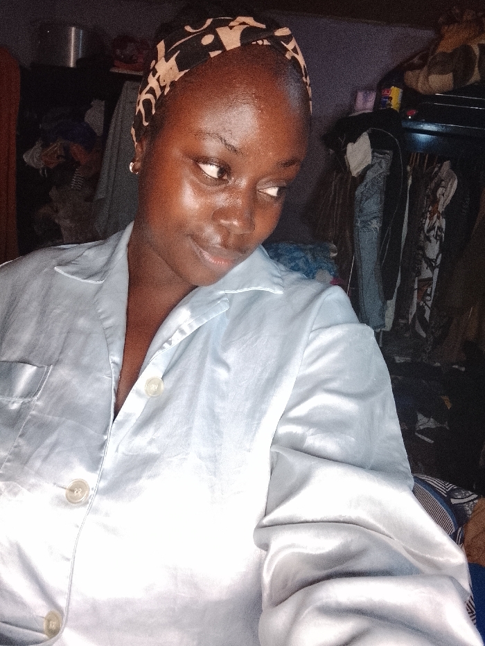 A young woman with smooth, dark skin and a patterned headband gazes to the right with a slight smile. She wears a light blue satin pajama top with three visible buttons. The background is cluttered with clothes hanging in a dark closet and various items stacked on shelves. The lighting is dim, creating a soft, slightly glossy effect on the woman's face and shirt.
