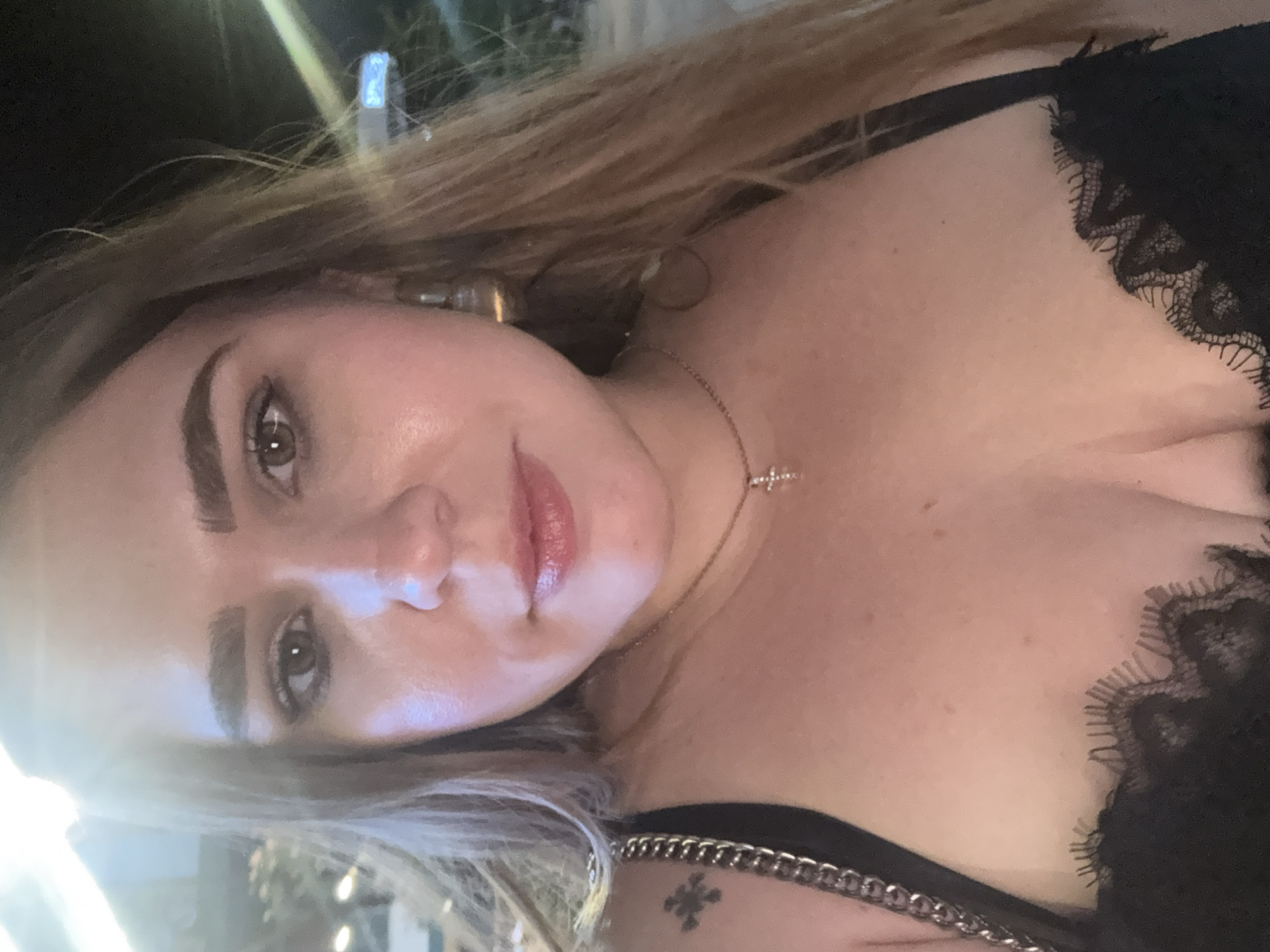 The media file is a close-up, selfie-style shot of a young woman with blonde hair. She is wearing a black lace top and a silver chain necklace with a small cross pendant. Her makeup is natural, with a focus on her eyes and lips. The background is dark and blurry, suggesting an evening setting, possibly outdoors with some ambient lighting. The overall mood appears relaxed and candid. There is no visible text in the image.