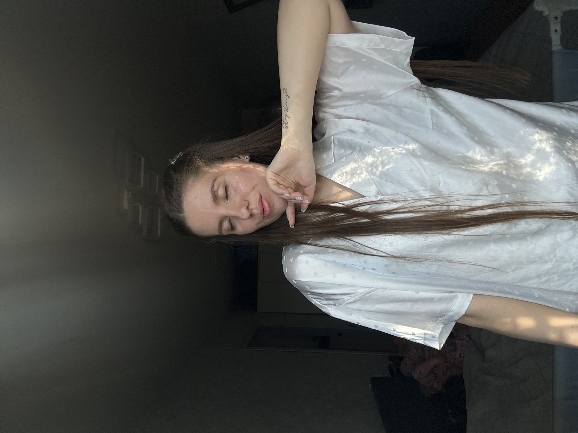 A woman with long brown hair poses in a white robe with a heart pattern. Her eyes are closed, and her hand is near her face. She has a tattoo on her arm. The background is dark, with hints of furniture and a window. The location is not discernible from the image.