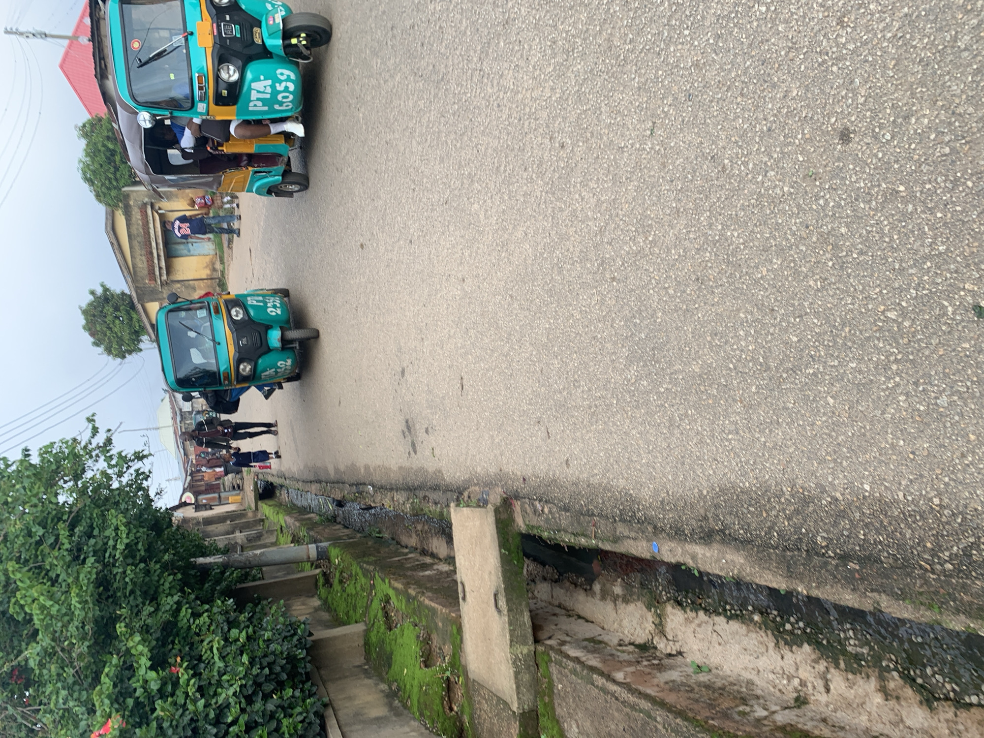 In the city of Jos, Nigeria, this image captures a street scene featuring two turquoise and yellow auto-rickshaws, also known as Keke Napeps, one of which is clearly labeled "PTA 6059." These three-wheeled vehicles, common modes of public transportation, are parked or moving on a paved road. To the left of the road, a concrete drainage channel runs alongside a weathered wall, with overgrown green foliage and moss indicating a humid environment. A concrete slab serves as a rudimentary bridge over the drain. In the background, several buildings with varying roof colors, including red and yellow, suggest a mix of residential and commercial structures. The sky is overcast, hinting at a potentially cloudy or rainy day. Pedestrians, including a man in a dark jacket and a child in a blue outfit, are visible walking along the side of the road. The overall impression is a snapshot of everyday life in a Nigerian urban setting, with its characteristic modes of transport and architectural styles.