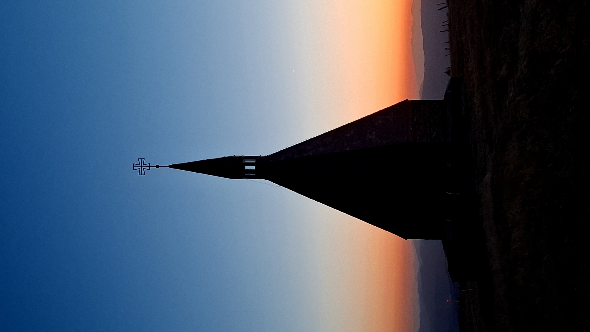 The image captures a silhouette of a chapel with a cross at its peak against a twilight sky. The sky transitions from a deep blue at the top to hues of orange and yellow near the horizon. In the foreground, a dark, silhouetted landscape with rolling hills and dry vegetation is visible. The chapel, with its pointed roof, is centrally placed and dominates the frame. The scene appears to be set in a remote, elevated location. Based on external information, this chapel is located in Waldbach-Mönichwald, Austria, and is known as the Waldbacher Mariahilfkapelle.