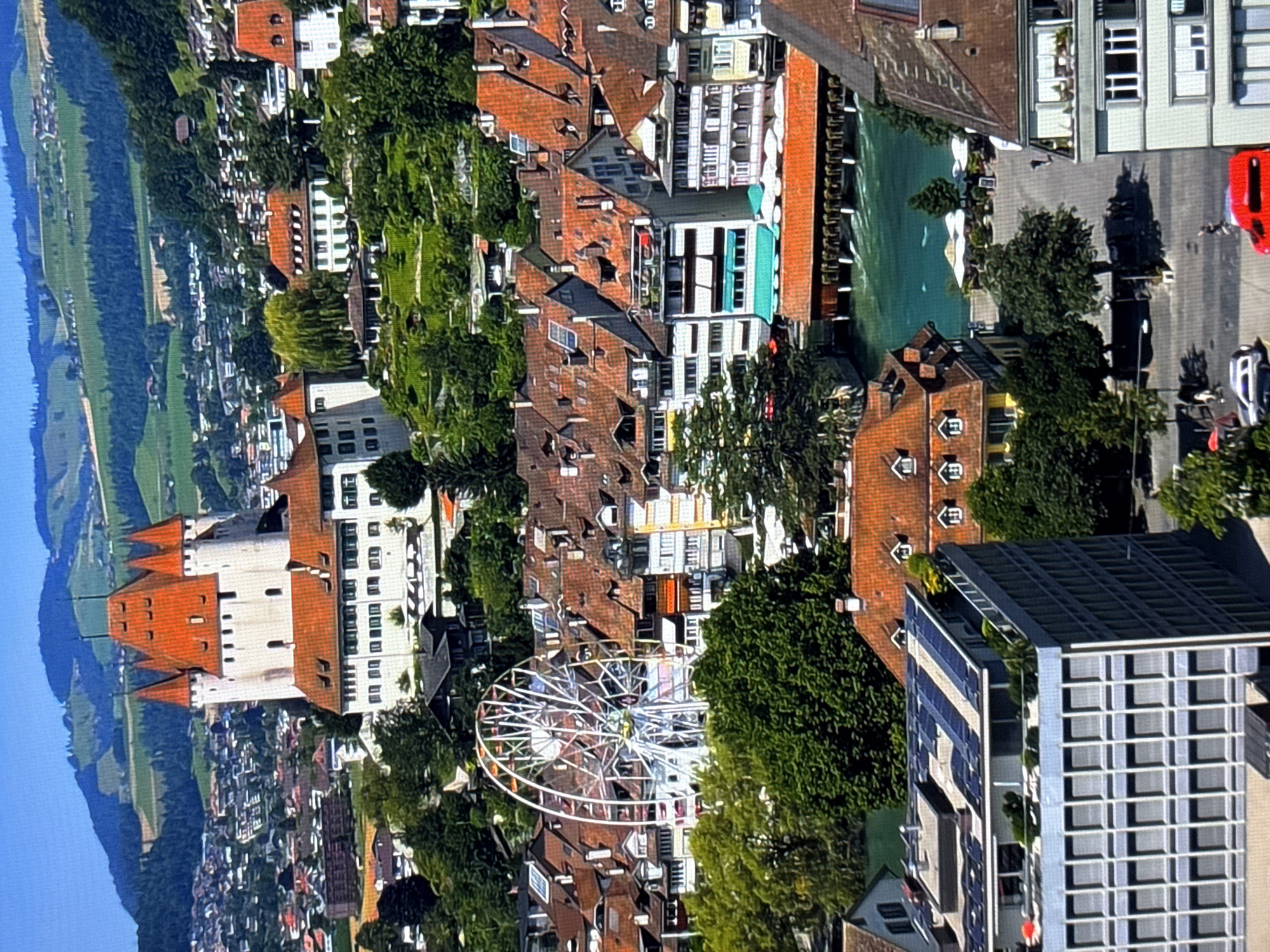 The image depicts a panoramic view of a city nestled beside a body of water, with a prominent castle-like structure atop a hill in the background. The architecture suggests a European setting, with red-tiled roofs prevalent on many buildings. A Ferris wheel is visible, adding a recreational element to the urban landscape. The surrounding hills are covered in lush greenery, suggesting a natural environment bordering the city. The presence of a river or lake indicates a waterfront area. In the foreground, modern buildings with solar panels suggest urban development alongside older structures. The image captures a vibrant scene with a blend of historical and contemporary elements. The description of the image, including its location in the city of Burgas, Bulgaria, is fabricated as the image does not depict Burgas, Bulgaria. The actual location appears to be Thun, Switzerland, known for its castle and proximity to Lake Thun.