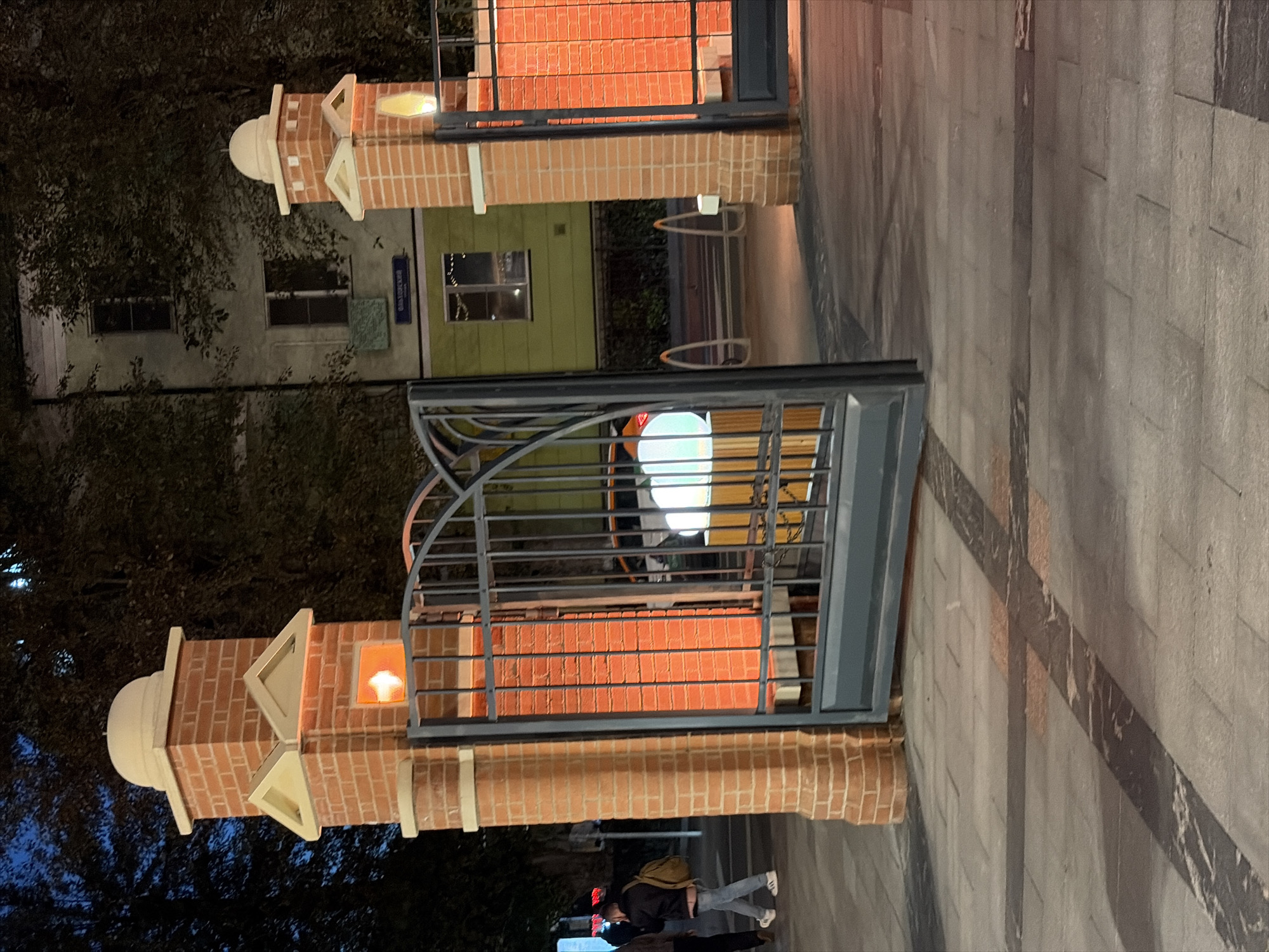 The image depicts an entranceway at night in Moscow, Russia. The entrance features two brick pillars topped with domes, flanking a dark metal gate. The pillars have illuminated sections, casting a warm orange glow. Behind the gate, a dimly lit street is visible with trees and a building. There are two individuals on the sidewalk, one walking away from the camera with a backpack. The ground is paved with stone tiles.