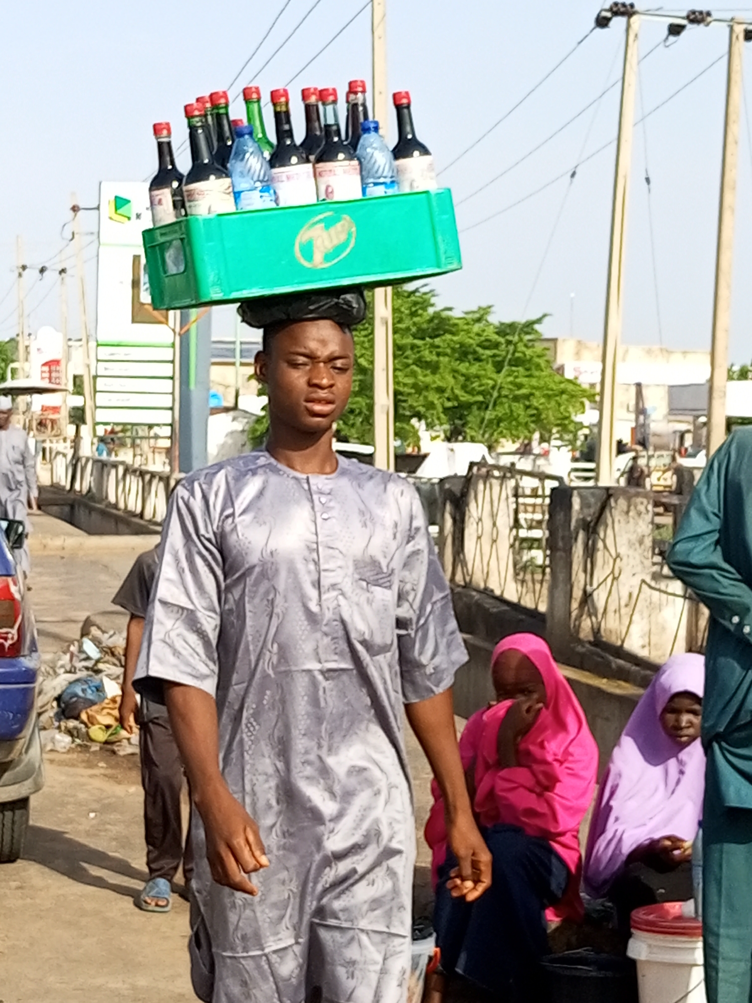 A young man, identified as a vendor, walks outdoors balancing a green plastic crate filled with bottled beverages on his head. He is wearing a traditional grey kaftan. Behind him, a few other people are visible, including two women wearing headscarves, one pink and one purple, sitting on a ledge. In the background, there are electric poles, trees, and buildings. To the left of the frame, part of a blue vehicle and some discarded waste are present on the ground. The scene appears to be a street in a city.
