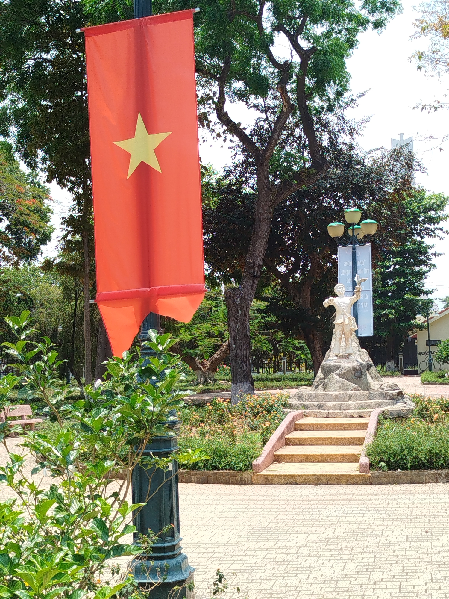 A vibrant photograph captures a scene in Liên Nghĩa, Vietnam, featuring a prominent Vietnamese flag hanging from a lamppost. The flag, a solid red with a five-pointed yellow star, is vertically oriented. The lamppost is dark green. Behind the flag, a statue of a young boy holding a bird is mounted atop stone steps. A street lamp with multiple globe-shaped lights stands behind the statue. Green trees provide a backdrop, and the ground is paved with light-colored bricks. Bushes with greenery are prominent in the foreground. A building with a white exterior is visible in the background, adding to the context of the scene.