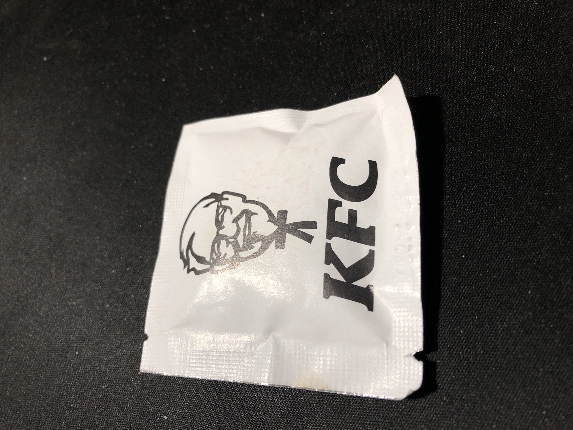 The image shows a close-up, overhead view of a single, white KFC sauce packet resting on a black, textured surface. The packet is oriented horizontally, with the KFC logo printed in black on the right side. The logo features the silhouette of Colonel Sanders on the left, followed by the letters "KFC" in a bold, sans-serif font. The packet appears to be made of a thin, crinkled material, with visible seams along the edges. The black surface beneath the packet has a subtle texture, possibly fabric or a coarse mat. There are no other objects or elements in the frame, and the lighting creates a slight shadow to the right of the packet, suggesting the light source is from the upper left. No specific time of day, weather, or location cues are discernible from the image itself, and no other text is visible.