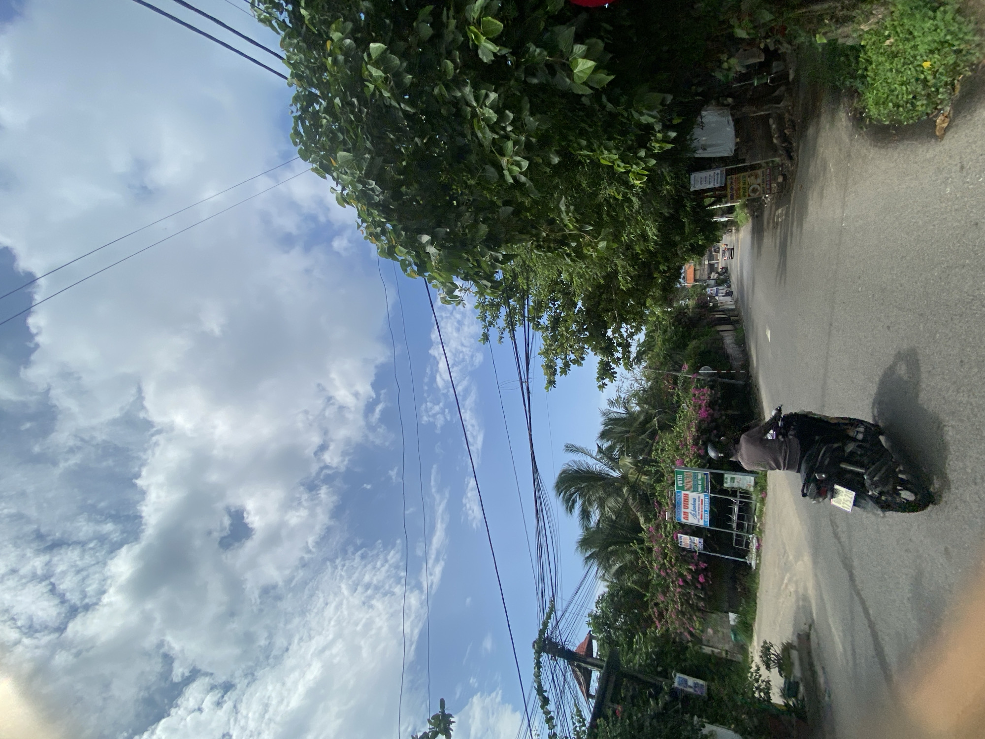 A single person is riding a motorcycle down a paved road in Mũi Né, Vietnam. The road curves to the right and is lined with trees and vegetation on both sides. Overhead, a tangle of electrical wires crisscrosses the bright blue sky, which is dotted with white, fluffy clouds. In the background, signs for businesses, including "AN VINH HOTEL" and "PHO NGON," are visible. A few buildings and palm trees can be seen further down the road. The motorcyclist is wearing a dark shirt and a helmet, and the motorcycle is black with a license plate. The scene appears to be in a semi-urban or tropical environment.