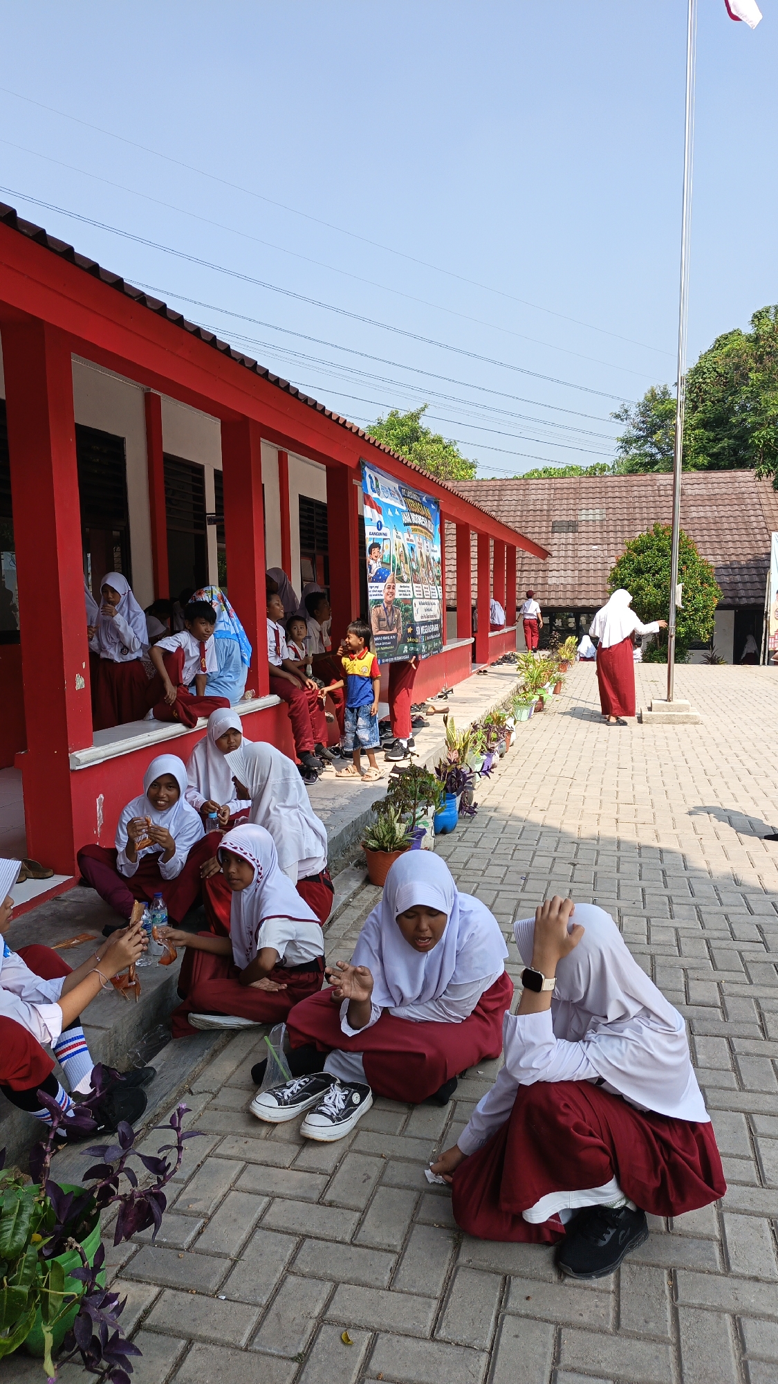 The image depicts a sunny outdoor scene at a school in Pasir Bolang, Indonesia, likely during a break or recess. Several young students, predominantly girls wearing white shirts, red skirts or maroons, and white hijabs, are seen sitting on a paved ground and on a raised platform. Some are engaged in eating or drinking, while others appear to be conversing. One student in the foreground is looking up and gesturing with her hands. In the background, a group of students are seated on a raised concrete area in front of a red-trimmed building with open doorways. A flag pole stands to the right of the image, and a banner with text and images is displayed on the building. The environment is characterized by a paved courtyard, potted plants, and a clear blue sky. The architecture suggests a typical Indonesian school setting.