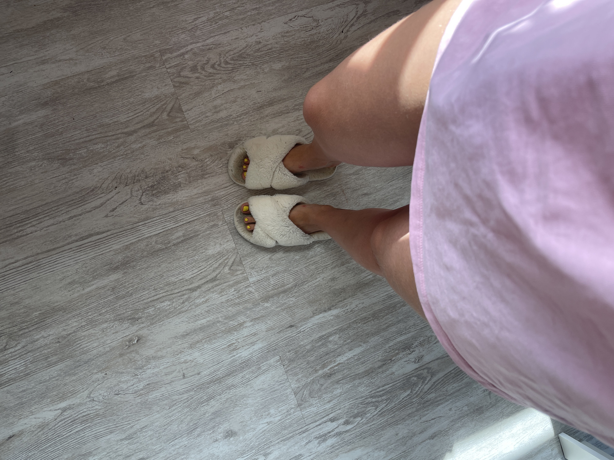 An overhead shot captures a person's legs and feet, adorned with fluffy white slippers and bright yellow nail polish, standing on a light-colored wooden floor. The floorboards are oriented diagonally, creating a dynamic visual texture. A soft pink garment, likely a dress or skirt, occupies the foreground, partially obscuring the lower torso and thighs. Sunlight casts a distinct shadow across the right leg and garment, indicating illumination from a window or light source to the left. The scene appears to be indoors, possibly in a residential setting within Мосметаллоконструкция Онт, Russia, characterized by the common flooring material and the casual attire.