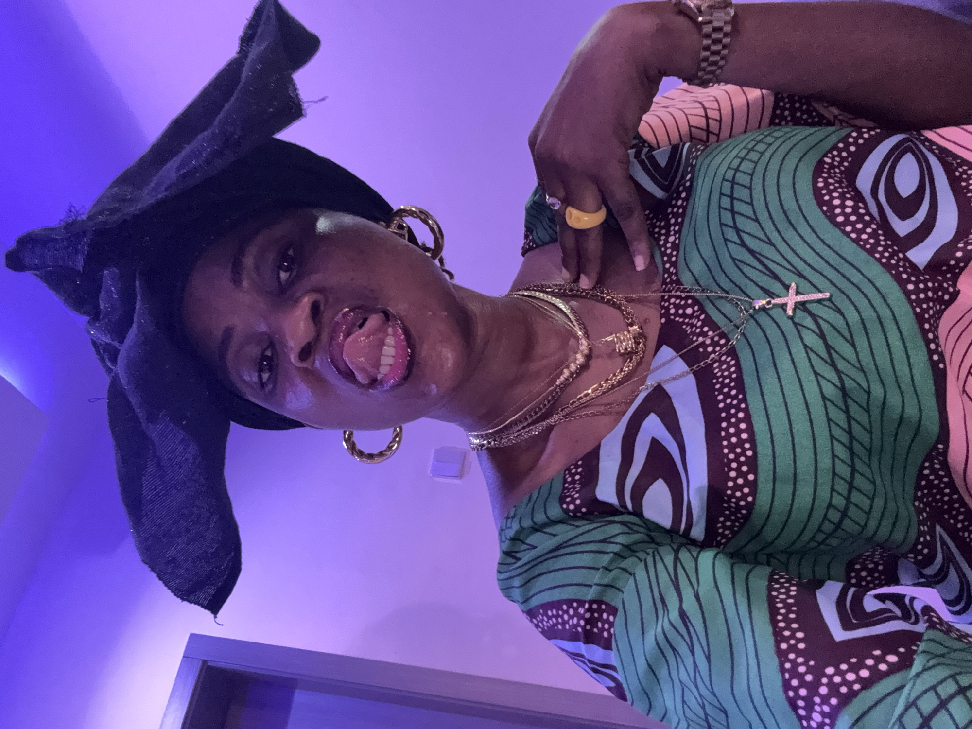 The image shows a medium shot of a woman with dark skin, wearing a black headwrap and a colorful patterned dress. She is sticking her tongue out, which has pink lipstick on it, and her mouth is slightly open. The woman is adorned with several gold necklaces, large hoop earrings, and rings on her fingers, one of which is bright yellow and oval-shaped. Her right hand is placed on her chest. She is also wearing a watch on her left wrist. The background is a solid purple wall with a subtle gradient, possibly due to artificial lighting. There is a doorframe visible on the left side of the image. The overall lighting is dim and illuminated by purple light, creating a somewhat dramatic atmosphere. The specific location in Ajara, Nigeria, is not identifiable from the visual cues present in the image.
