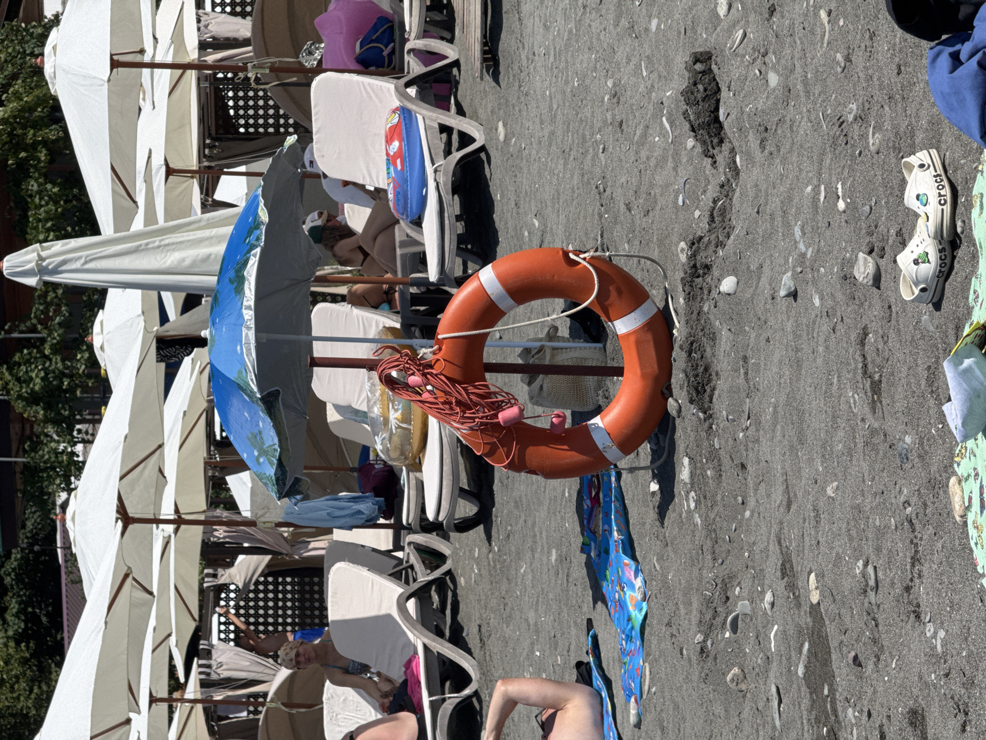 The image depicts a beach scene in Sochi, Russia, with many beach umbrellas set up over lounge chairs. In the foreground, a bright orange life preserver with white stripes is propped up on a stand. A blue and white beach towel lies on the dark, pebbly sand to the left of the life preserver. Further back, several people are visible lounging on chairs under the umbrellas. One person is partially visible on a lounge chair in the left foreground, wearing a swimsuit and a headscarf. Another person in a swimsuit is seated on a lounge chair in the background. A blue inflatable ring is placed on a lounge chair behind the life preserver. A few more lounge chairs and umbrellas are visible stretching into the background, suggesting a popular beach resort. In the bottom right corner, a pair of white Crocs shoes is placed on the sand. The overall atmosphere is one of a sunny day at the beach.