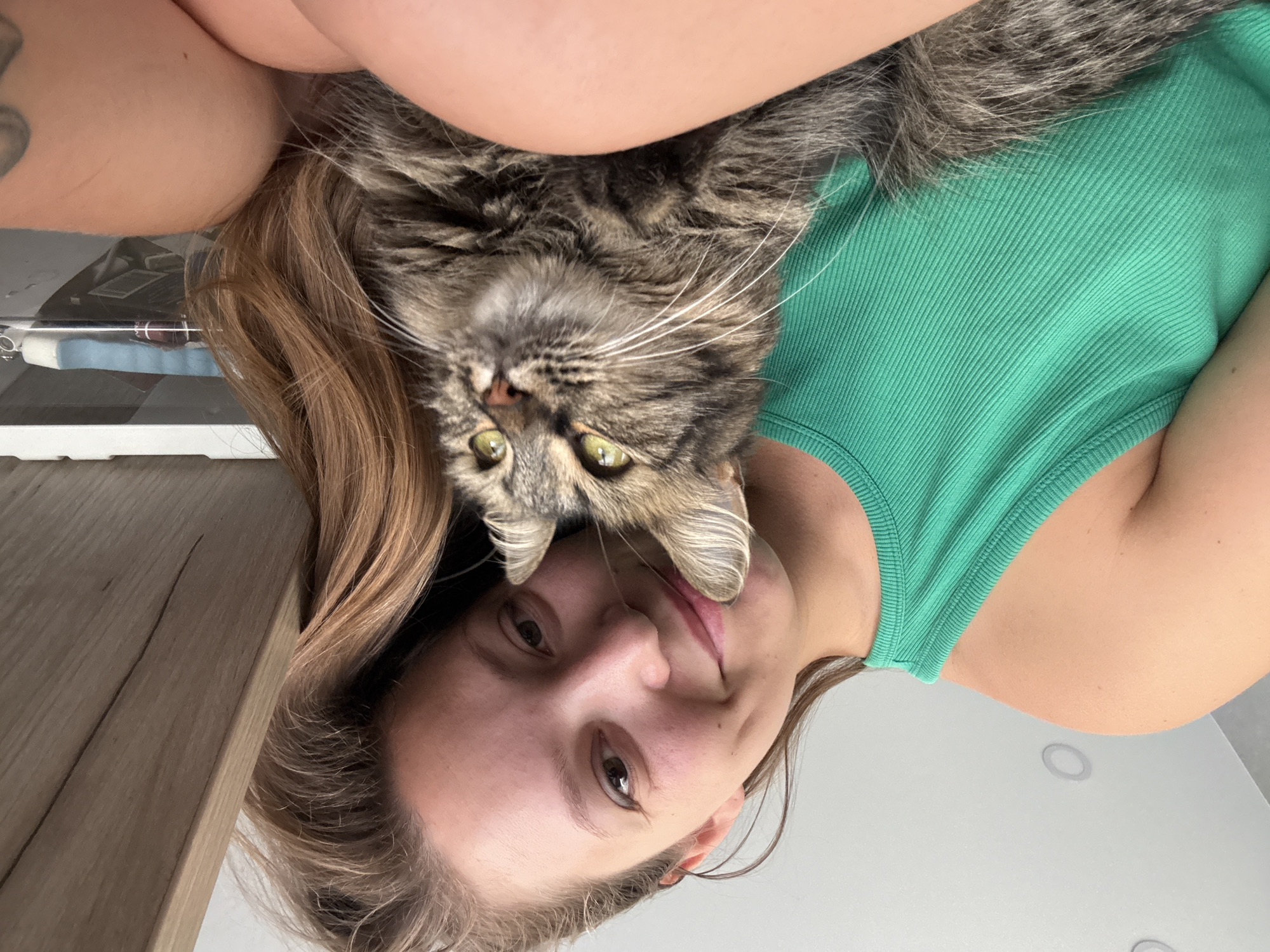 In this close-up selfie, a young woman with long, brown hair and a bright green tank top is holding a tabby cat. The woman is smiling and leaning her head against the cat, who is looking away from the camera. The image appears to be taken indoors, possibly in a home. The background is out of focus, but it seems to include a white ceiling with recessed lighting and a wooden surface.