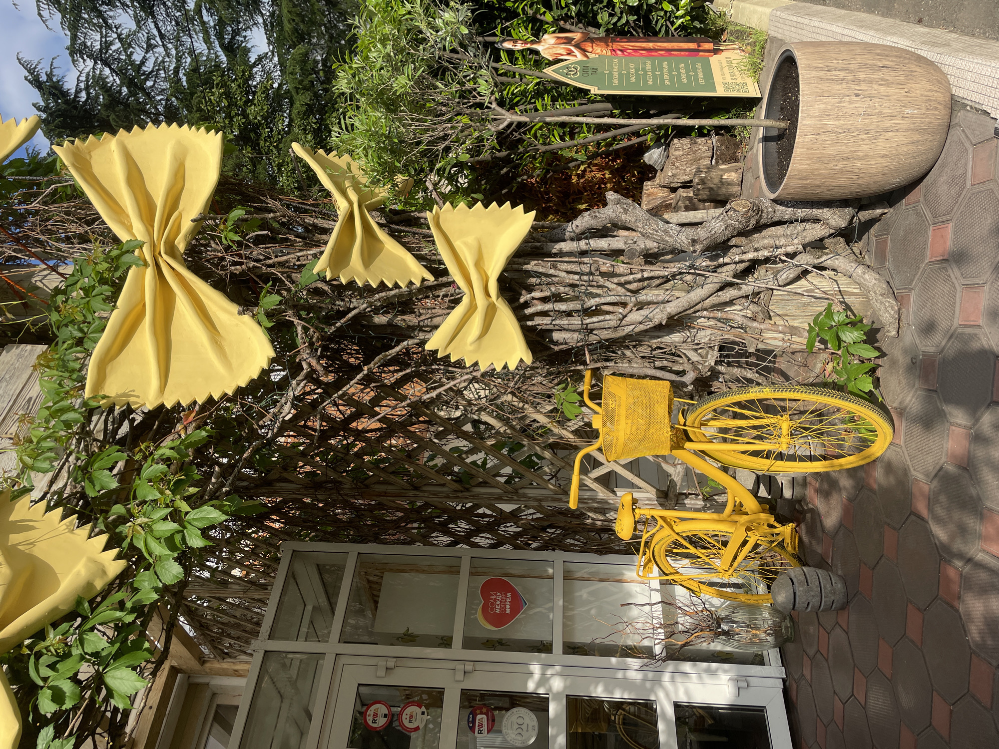 The image captures an outdoor scene, likely a decorative display in Sochi, Russia. Prominently featured are three large, yellow pasta-shaped decorations (farfalle) affixed to a lattice structure intertwined with branches and greenery. A yellow bicycle is positioned against the lattice, appearing to be part of the display.
Below the bicycle, a glass-paned doorway is visible, indicating a building entrance. Stickers and a heart-shaped graphic with Cyrillic text ("СОЧИ МЕЖДУ СОНЦЕМ МОРЕМ") are visible on the glass.
To the right of the pasta and bicycle arrangement, a tree trunk-shaped planter sits atop a collection of branches. Behind this, a sign with text and an image of a woman provides information in Russian about "Thai City" spa services. The ground is paved with red and brown hexagonal tiles. The overall scene has a whimsical and artistic feel.