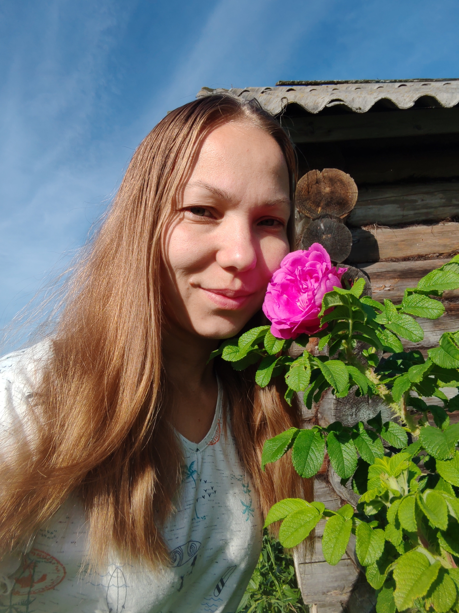 The image shows a young woman with long, reddish-brown hair, taking a selfie outdoors. She is smiling gently and has a pink rose near her face. She is wearing a light-colored t-shirt with a pattern of palm trees and surfboards. Behind her is a rustic wooden building, possibly a cabin, constructed from logs. The roof of the building appears to be made of corrugated material. The sky is bright blue with some wispy clouds. Lush green foliage, including the rose bush, is visible in the foreground and around the building. The overall impression is one of a sunny day in a rural or semi-rural setting, potentially a dacha or a village house in the vicinity of Чухлэм, Russia, given the context.
