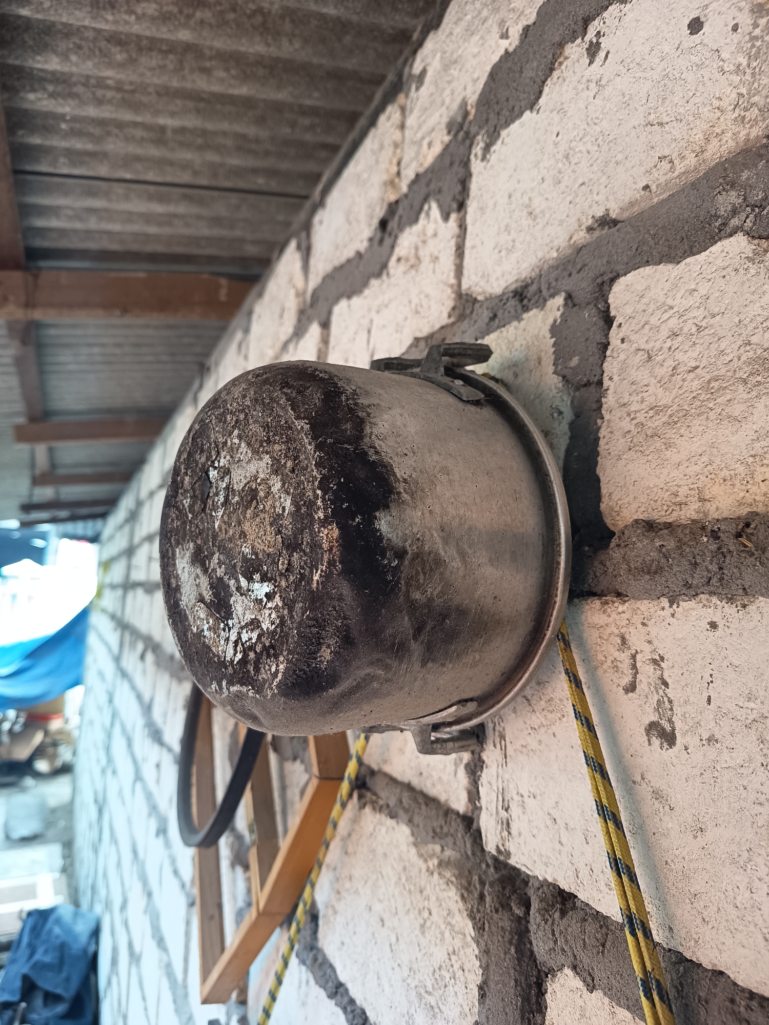 A pot with black soot is attached to a white brick wall. A yellow and black rope is hanging from the pot. A wooden frame is also attached to the wall behind the pot. The wall is under a corrugated roof. The image appears to be taken in an outdoor area, possibly a construction site or a workshop, in Sugio, Indonesia.