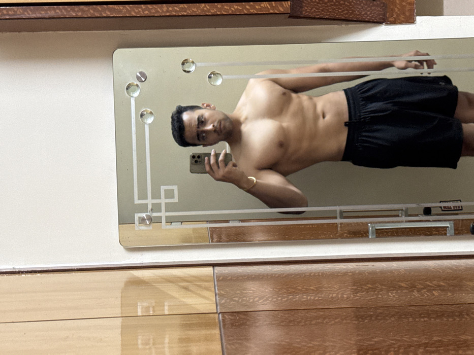A man is reflected in a mirror. He is shirtless, wearing black shorts. He is holding a cell phone in his hand, presumably taking a photo. He is also wearing a gold bracelet on his left wrist. The mirror has a decorative border and is mounted on a wall with wooden paneling below. The man's face is visible in the reflection.