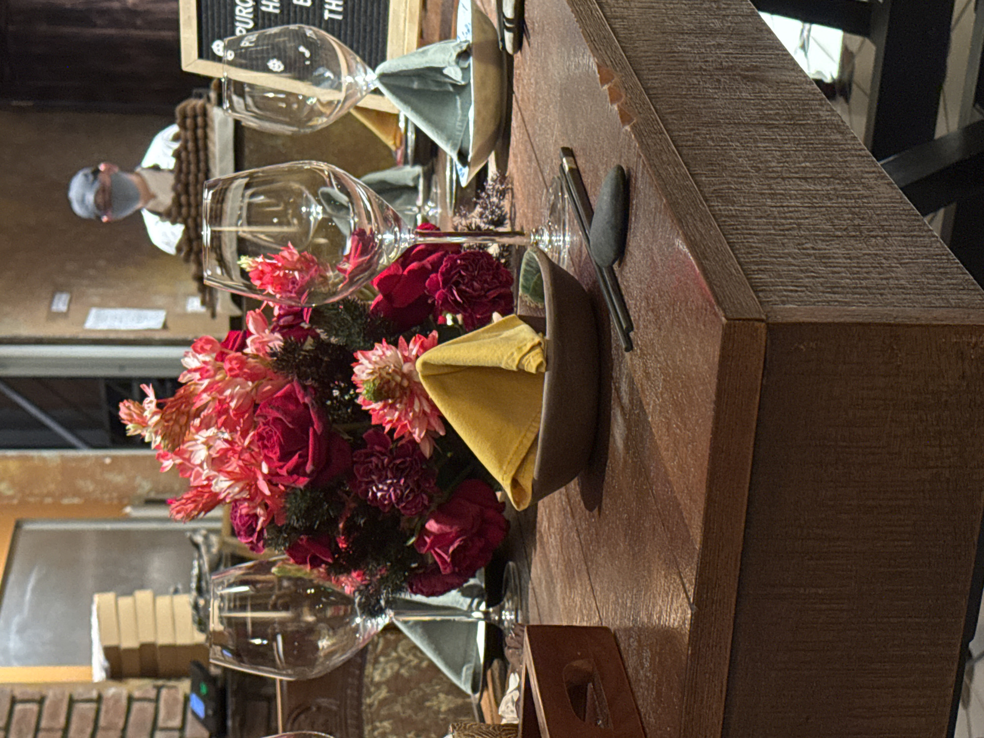 The image depicts a restaurant setting in Hà Nội, Vietnam. A wooden table is adorned with a floral centerpiece composed of red and pink flowers, set in a glass vase. Place settings are present, featuring napkins folded in small bowls alongside silverware. Two wine glasses sit nearby, and another table setting can be partially seen in the background. A person wearing a face mask and a white shirt is also visible in the background. The restaurant's interior is dimly lit with some natural light coming through the windows.