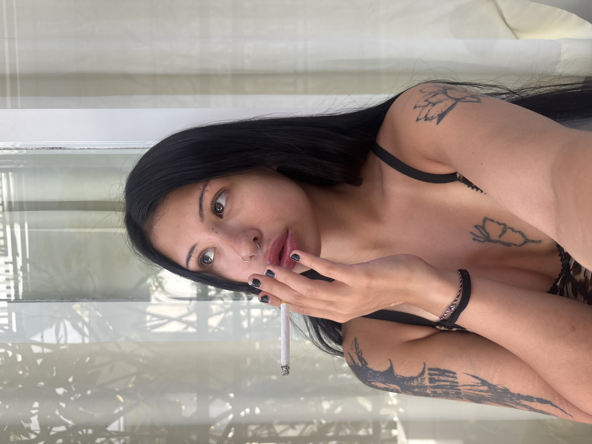 A young woman with long, dark hair and tattoos on her arms and chest is smoking a cigarette and looking upwards. She has a nose ring and dark nail polish. She is wearing a dark bra. The background is blurred and appears to be an indoor setting with sheer curtains and sunlight streaming in.