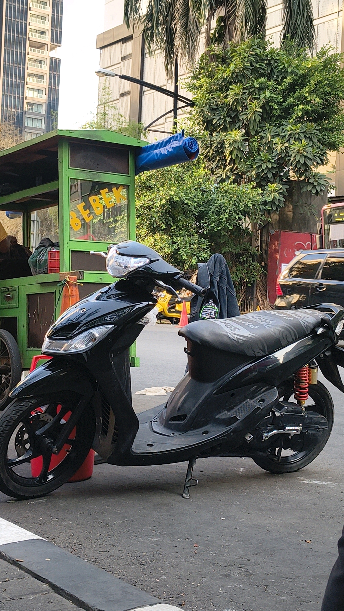 A black scooter is parked on an asphalt street in front of a green food cart in a city. The food cart has a sign that reads "BEBEK" in yellow letters and a blue rolled-up tarp attached to its roof. A black denim jacket is draped over the back of the scooter's seat. In the background, there are buildings, trees, and other vehicles, including a black car and a yellow vehicle. The scene appears to be set in South Jakarta, Indonesia, given the architectural style of the buildings and the presence of a food cart selling "bebek" (duck), which is a popular dish in Indonesia. The lighting suggests it is daytime, possibly late afternoon.