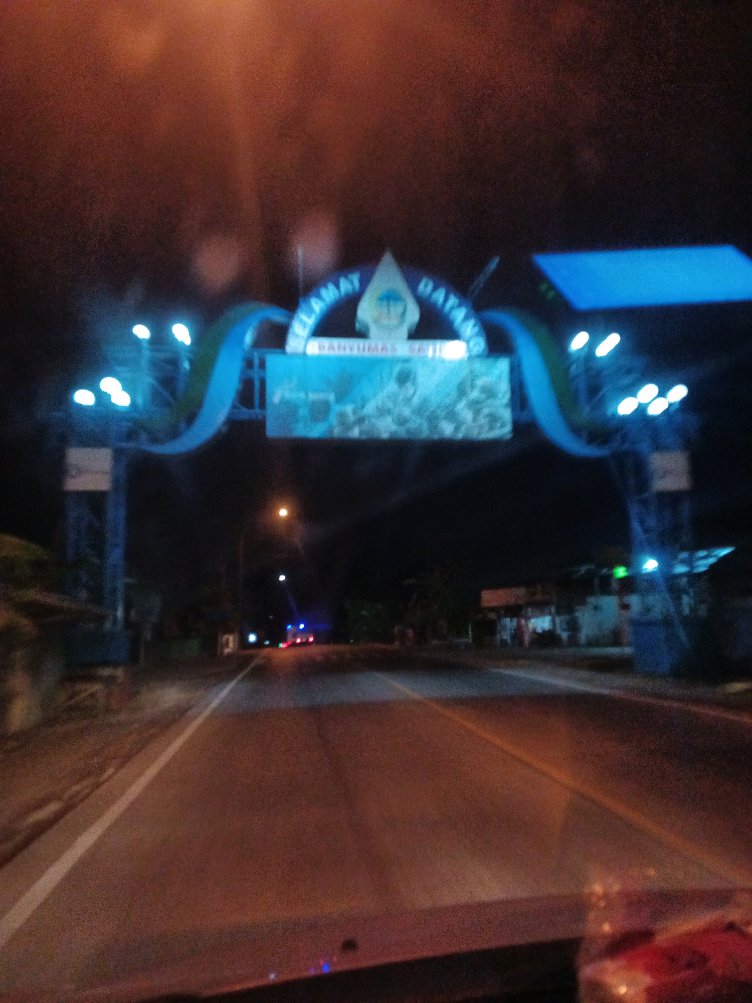 This is a photograph taken at night, from inside a vehicle, of a welcome gate arching over a road. The gate is illuminated with blue lights and features a sign with text that is partially obscured. The text appears to include "SELAMAT DATANG" and "BANYUMAS SATU". There is a graphic on the sign that resembles a traditional Indonesian headdress. The road in front of the gate is paved and has a white line marking the lane. The surrounding environment is dark, with some trees and buildings visible in the background. There are also streetlights and other vehicle lights in the distance. The photograph is somewhat blurry, suggesting it was taken while the vehicle was in motion.