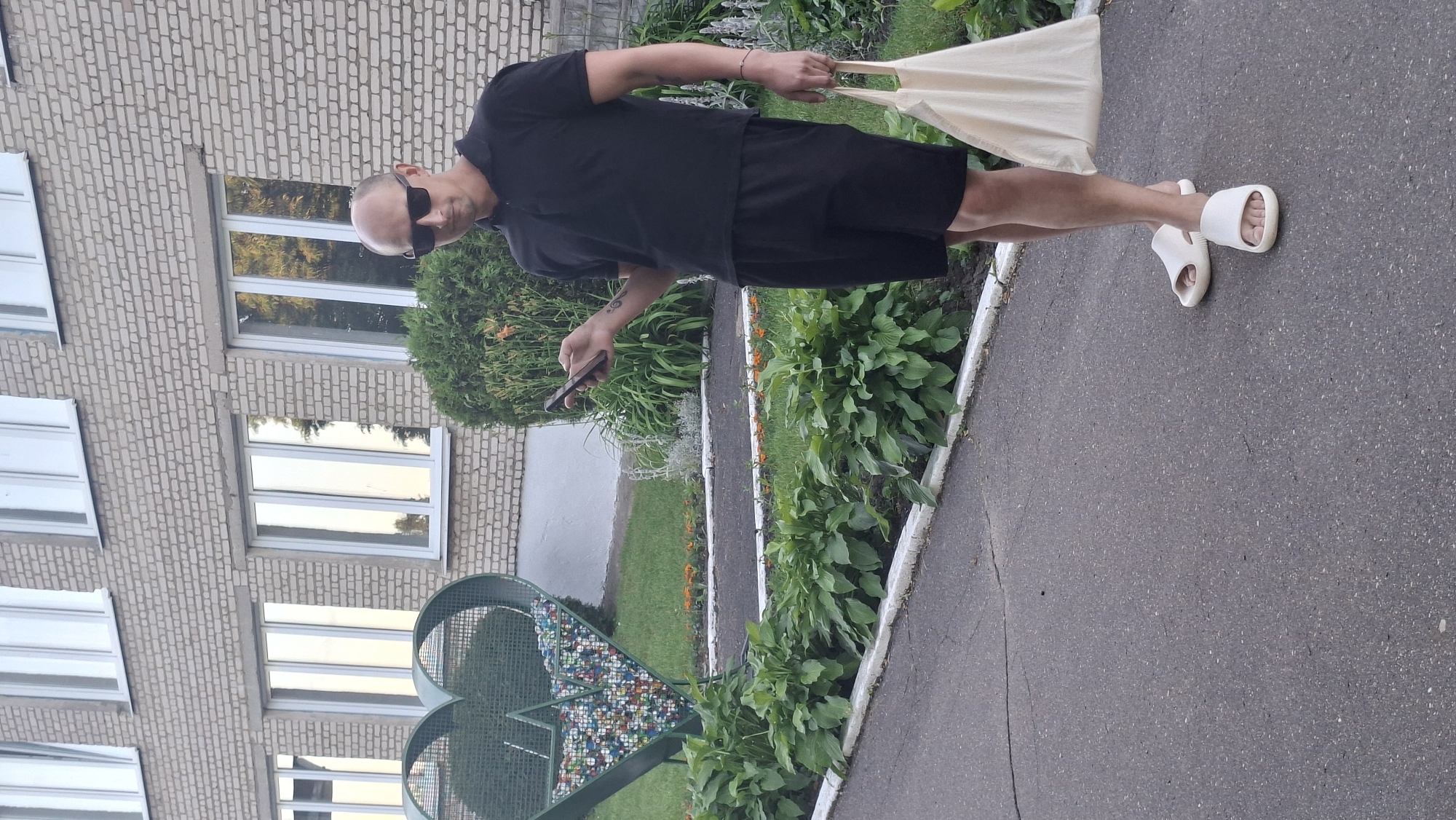A bald man wearing sunglasses, a black polo shirt, and black shorts stands on a paved walkway in front of a brick building. He is holding a smartphone in his left hand and a light-colored tote bag in his right. To his left is a large, green, heart-shaped trash receptacle filled with colorful objects. The building has multiple windows with reflective glass. Behind the trash receptacle and to the right of the man, there are various green plants and some orange flowers. The scene appears to be outdoors in an urban or suburban setting.
