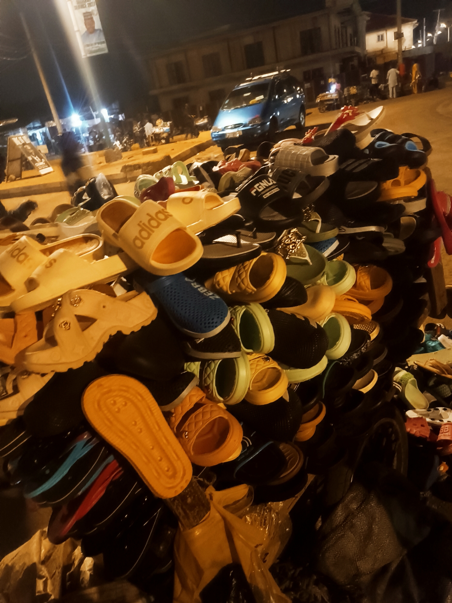 At night in Bukuru, Nigeria, a pile of colorful sandals, slides, and flip-flops sit on a surface outdoors. The shoes are mostly plastic and rubber. In the background, there are people, buildings, a blue car, and a banner with a portrait on a pole. There are some tables and signs to the left, also people standing around.