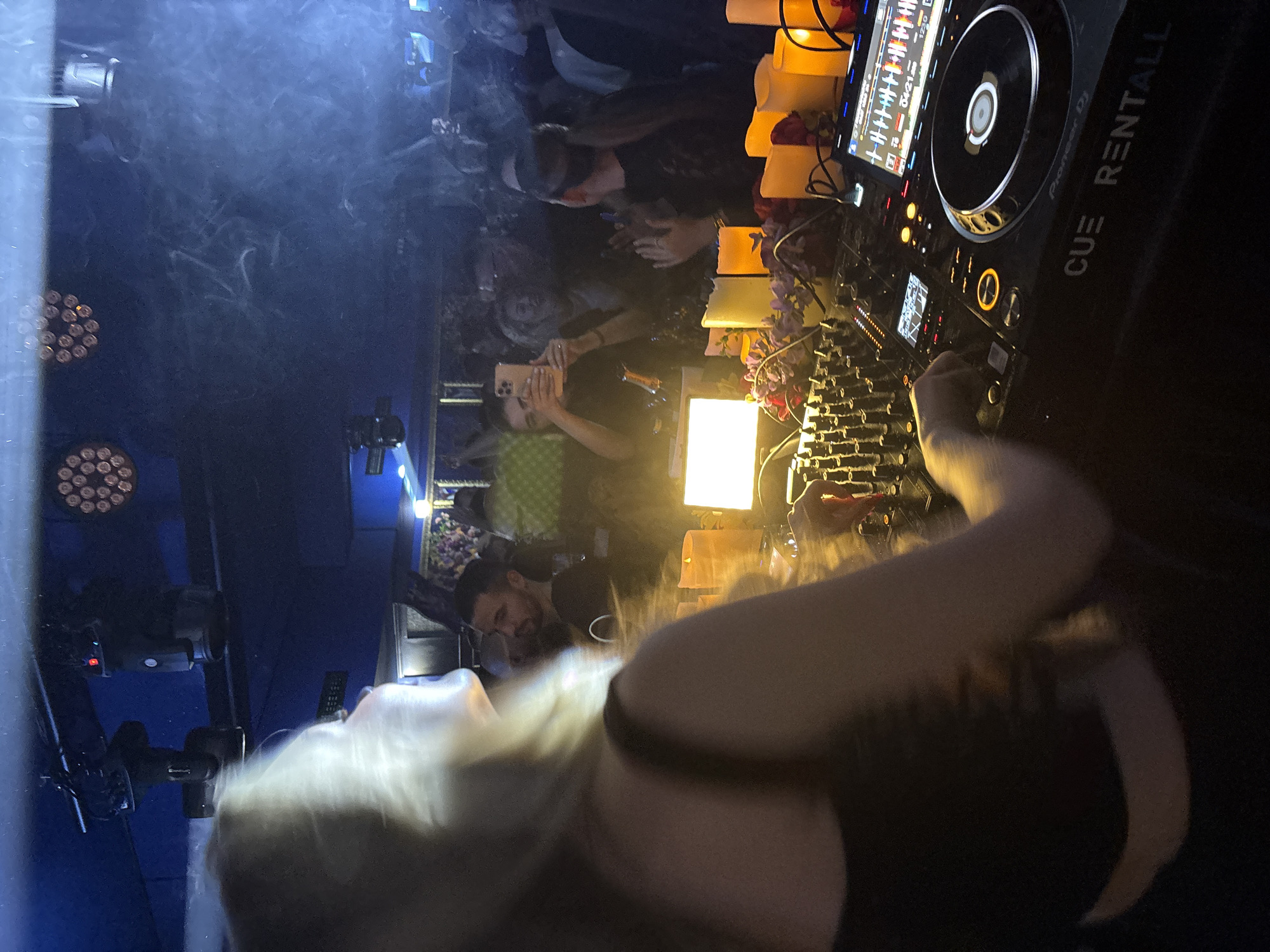 The image depicts a DJ performing in a nightclub setting. The foreground features the DJ equipment, including a Pioneer DJ setup with a mixer, turntables, and a display screen. The DJ is a fair-skinned woman with long blonde hair wearing a black tank top. Artificial candles and floral arrangements decorate the DJ booth.
In the background, there is a crowd of people, some holding up their phones. The nightclub has blue lighting and a haze or smoke effect in the air. The crowd appears to be in a dimly lit environment.
The city and country are Sofia, Bulgaria.