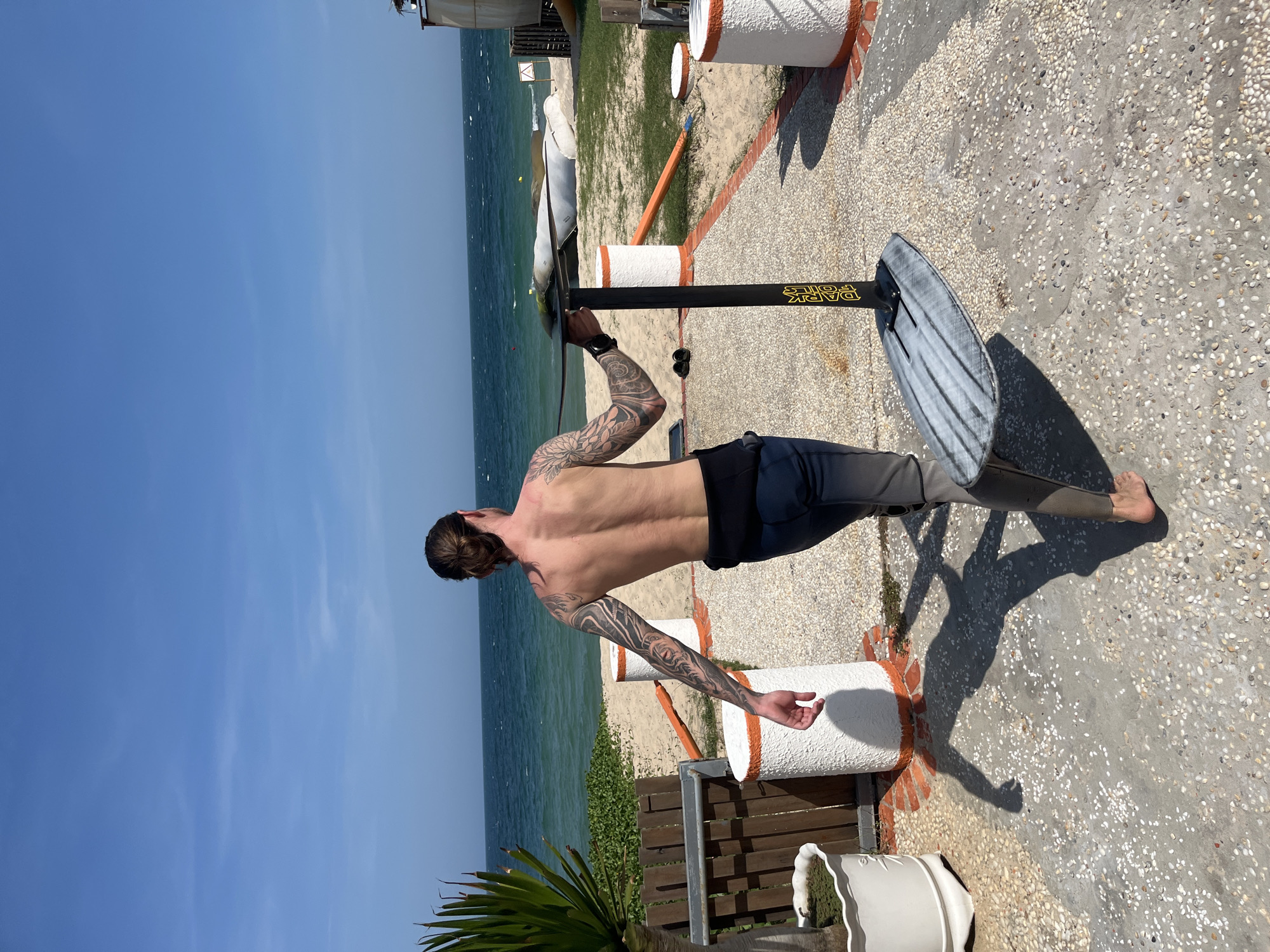 A shirtless man with dark hair pulled back in a bun and extensive arm and back tattoos walks away from the camera on a sunny day. He is wearing dark water sports pants. He carries a hydrofoil board, which consists of a grey, elongated board attached to a vertical black mast with the words "DARK FOILS" in gold lettering. The man is walking on a paved pathway composed of small, multicolored pebbles mixed with concrete, bordering a sandy beach. In the background, the turquoise ocean meets a clear blue sky with a few wispy clouds. A section of beach is visible with green foliage and some scattered white and orange cylindrical objects along the pathway. Further back on the beach, several boats are pulled up. The scene suggests an outdoor recreational area, likely near the coast of Phu Thuy Ward, Vietnam, suitable for water sports.