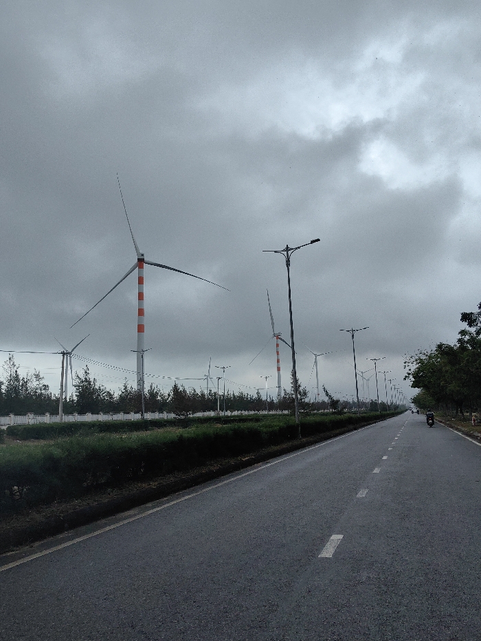 The image displays a straight asphalt road under an overcast, grey sky in East Quy Nhon Ward, Vietnam. On the left, the road is bordered by a median strip containing green shrubbery. Beyond this strip, a series of wind turbines with three blades each are visible; several towers feature prominent white and orange horizontal stripes, while others are entirely white. A white concrete barrier and rows of small trees separate the wind farm area from the roadside. Tall, dark streetlights with dual lamp heads line the left side of the road, extending into the background. The road itself features white dashed lane markings. On the right side of the road, dense green trees and vegetation form a boundary. In the distance, a single individual is observed operating a motorcycle, traveling away from the camera.
