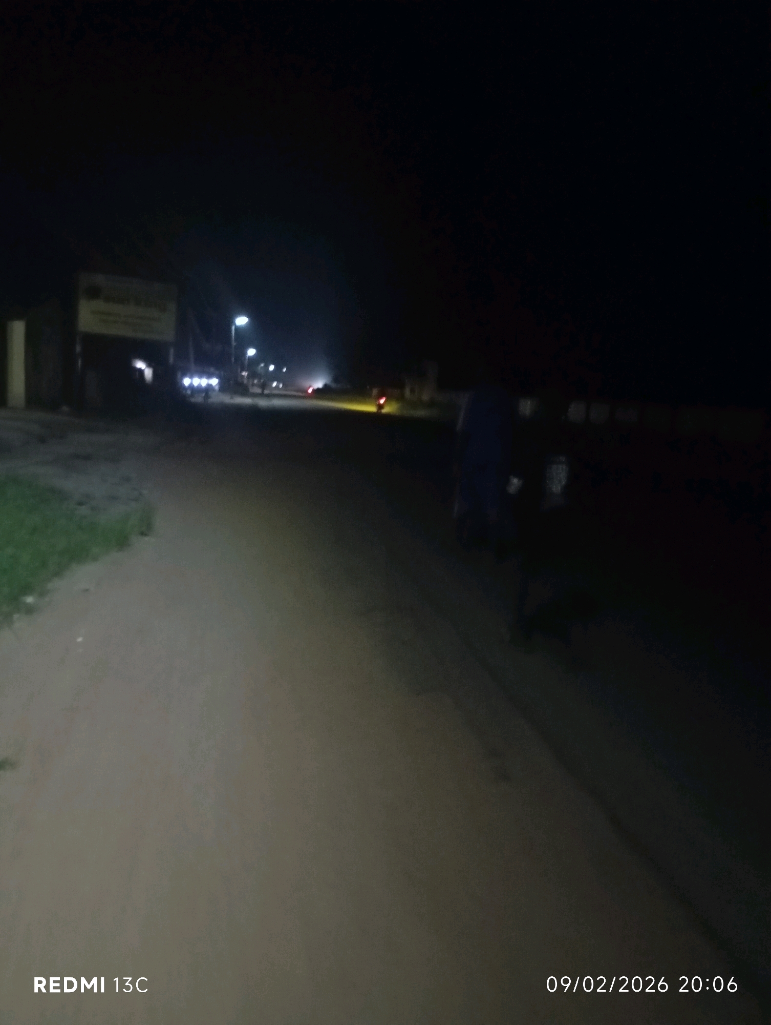 The image is a low-light, night-time photograph taken on a road in Shendam, Nigeria. The scene depicts a dirt road with a grassy verge on the left. In the distance, there are streetlights illuminating the road, and several vehicles are visible, including what appears to be a motorcycle with its headlights on. A sign is present on the left side of the road, though it is difficult to read due to the low light. The overall impression is of a quiet, rural road at night.
